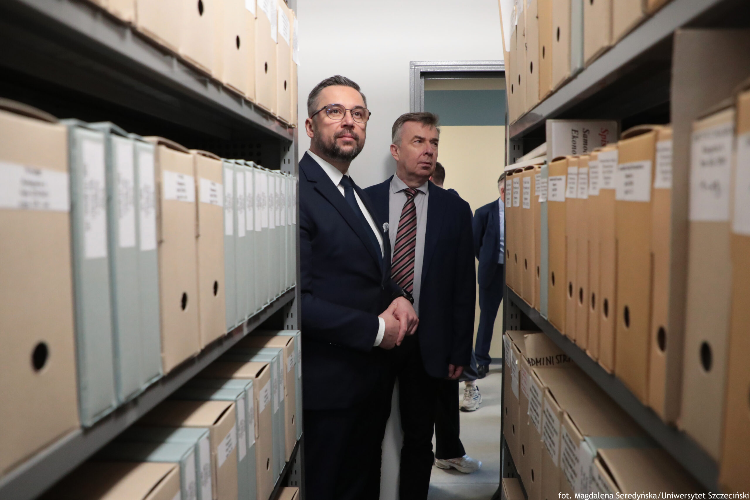 Korytarz między dwoma regałami archiwalnymi wypełnionymi teczkami i kartonowymi pudełkami z etykietami. Po obu stronach kadru stoją wysokie metalowe półki; na półkach równo ustawione jasnobeżowe teczki i pudła, część z okrągłymi otworami na grzbiecie. W środku przejścia stoją dwaj mężczyźni w garniturach. Mężczyzna po lewej ma ciemne włosy z siwizną, krótko przycięte, brodę i wąsy, okulary oraz ciemny garnitur z białą koszulą i krawatem; patrzy w lewą stronę kadru. Mężczyzna po prawej ma krótkie jasnobrązowe włosy, jest w ciemnym garniturze, białej koszuli i krawacie w ukośne pasy; również patrzy w lewą stronę kadru. W tle, przy otwartych drzwiach, widać fragmenty kolejnych osób, w tym nogi w jasnych spodniach i białych butach sportowych. W prawym dolnym rogu jest podpis: „fot. Magdalena Seredyńska/Uniwersytet Szczeciński”.