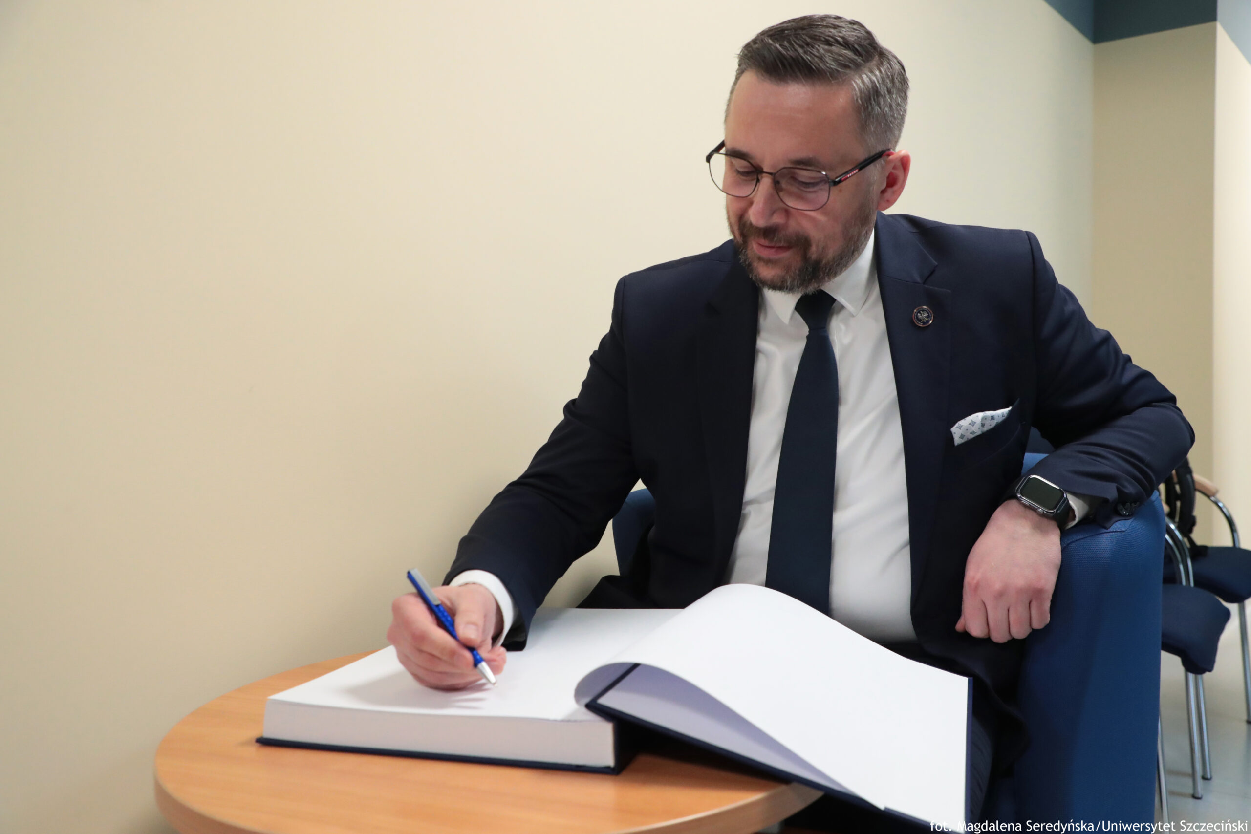 Mężczyzna w garniturze siedzący na niebieskim fotelu i podpisujący dokument w dużej księdze na okrągłym drewnianym stoliku. Ma krótkie ciemne włosy z siwizną, okulary i krótko przystrzyżoną brodę. Ubrany jest w ciemny garnitur, białą koszulę i ciemny krawat; w brustaszy ma jasną poszetkę, a na lewej ręce smartwatch. Trzyma niebieski długopis nad otwartą księgą z białymi stronami i ciemną okładką. Tło stanowi jasna ściana; z prawej strony widać fragment dodatkowych krzeseł. W prawym dolnym rogu jest mały napis: „fot. Magdalena Seredyńska / Uniwersytet Szczeciński”.
