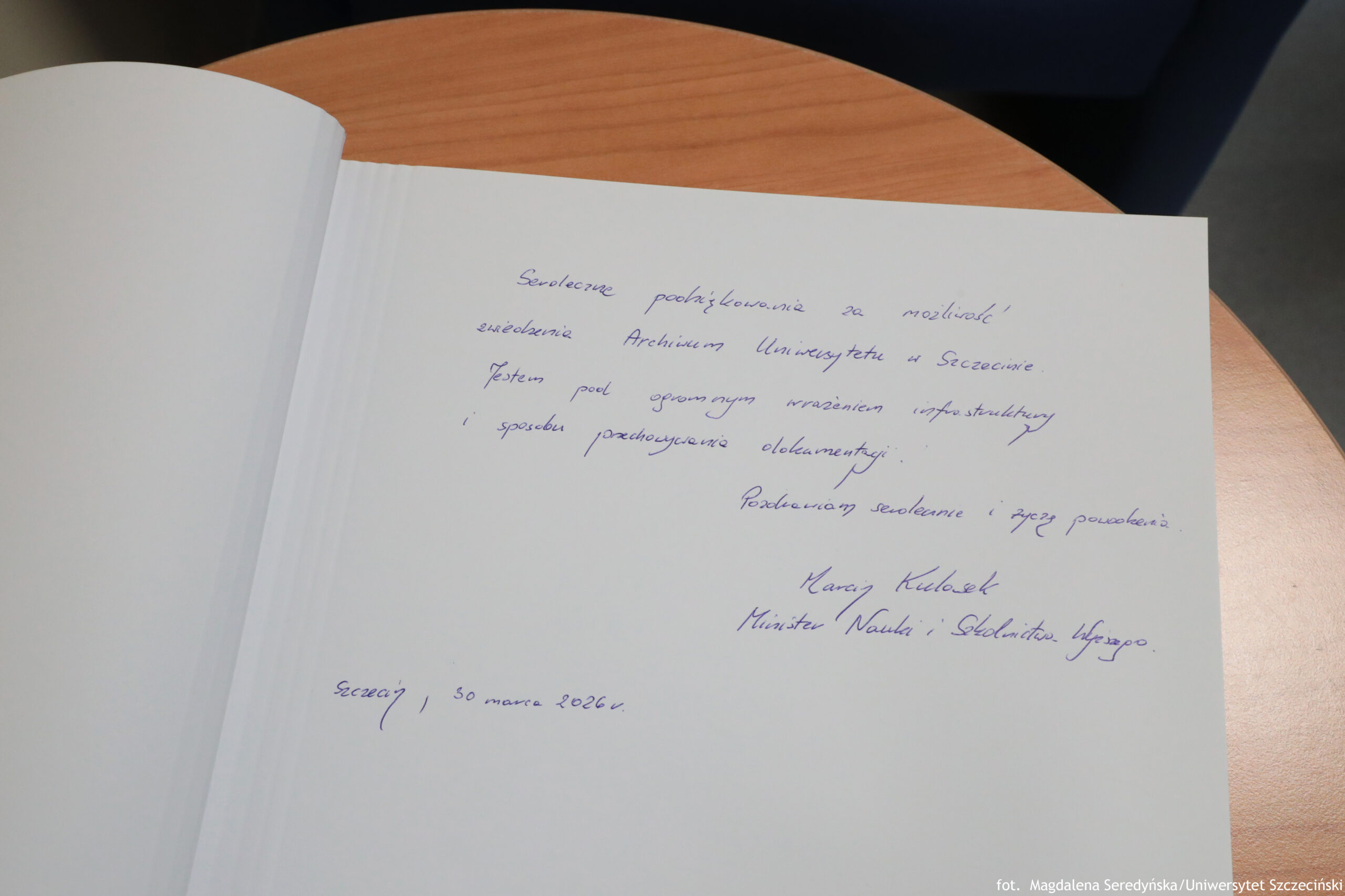 Otwarta księga pamiątkowa na drewnianym, okrągłym stole, z wpisem odręcznym niebieskim długopisem na prawej stronie. Wpis jest po polsku, po lewej stronie widać pustą stronę. Transkrypcja widocznego tekstu (pisownia jak na zdjęciu, miejscami słabo czytelna): „Serdeczne podziękowanie za możliwość zwiedzenia Archiwum Uniwersytetu w Szczecinie. Jestem pod ogromnym wrażeniem infrastruktury i sposobu przechowywania dokumentacji. Pozdrawiam serdecznie i życzę powodzenia Karol Kulasek Minister Nauki i Szkolnictwa Wyższego. Szczecin, 30 marca 2026 r.” W prawym dolnym rogu zdjęcia jest mały podpis: „fot. Magdalena Seredyńska/Uniwersytet Szczeciński”.