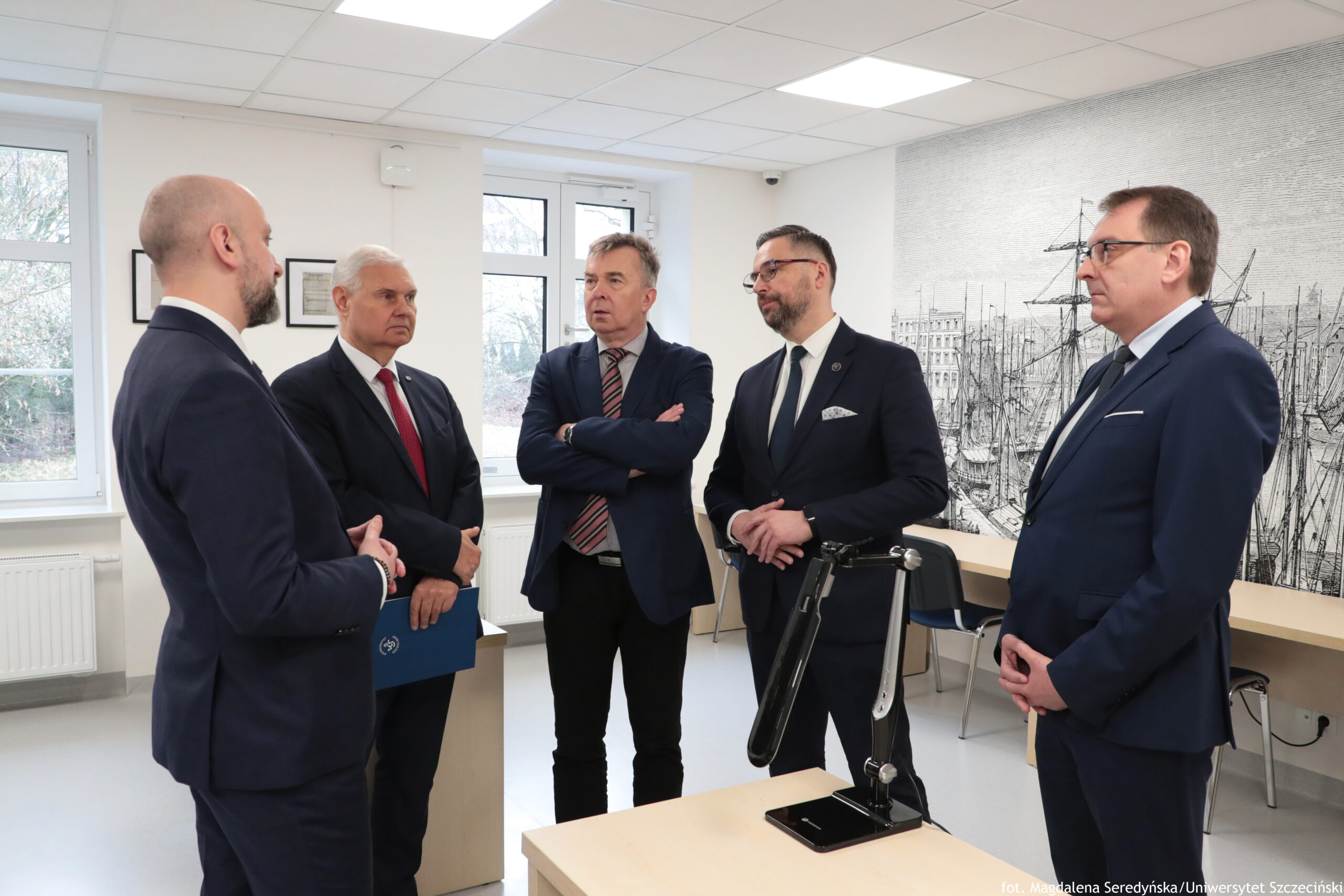 Grupa pięciu mężczyzn w garniturach stojących w jasnym pomieszczeniu biurowo-szkolnym i rozmawiających. Wszyscy są dorośli, w wieku mniej więcej od około 30 do 60+. Po lewej stronie stoi mężczyzna z ogoloną głową i krótką brodą, w ciemnym garniturze; jest ustawiony bokiem i zwrócony w stronę pozostałych. Obok niego stoi starszy mężczyzna z siwymi włosami, w ciemnym garniturze i czerwonym krawacie; trzyma niebieską teczkę. W środku stoi mężczyzna w ciemnym garniturze, z krótkimi siwymi włosami, z założonymi rękami i krawatem w ukośne pasy. Po jego prawej stronie stoi mężczyzna z ciemnymi włosami i brodą, w okularach, w ciemnym garniturze z białą poszetką. Najbardziej po prawej stoi mężczyzna w okularach, w ciemnym garniturze; ma ręce złożone z przodu. Pomieszczenie ma białe ściany i sufit z panelami oświetleniowymi. Z tyłu są duże okna. Po prawej stronie na ścianie jest duża grafika przedstawiająca port z żaglowcami (czarno-biała kreska). Wzdłuż ściany stoi długi blat/biurko i kilka krzeseł. Na pierwszym planie jest biurko z czarną lampką biurkową na podstawie. W prawym dolnym rogu widoczny jest podpis: „fot. Magdalena Seredyńska/Uniwersytet Szczeciński”.