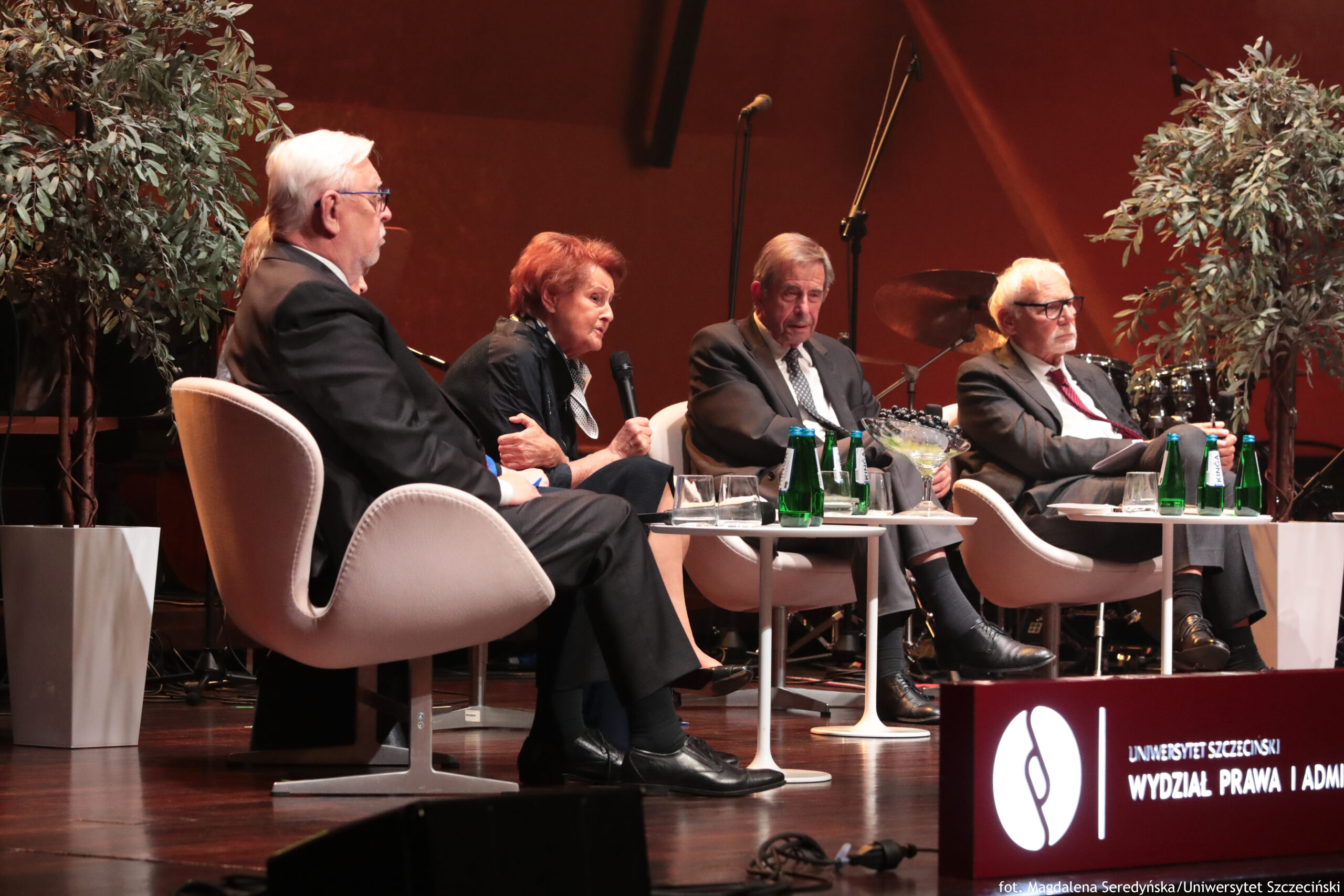 Scena. Ujęcie bokiem. Trzy okrągłe białe stoliki i cztery białe fotele, na których siedzą uczestnicy debaty (3 mężczyzn i 1 kobieta – w środku). Po bokach drzewka w wysokich białych doniczkach. W tle perkusja i mikrofony.