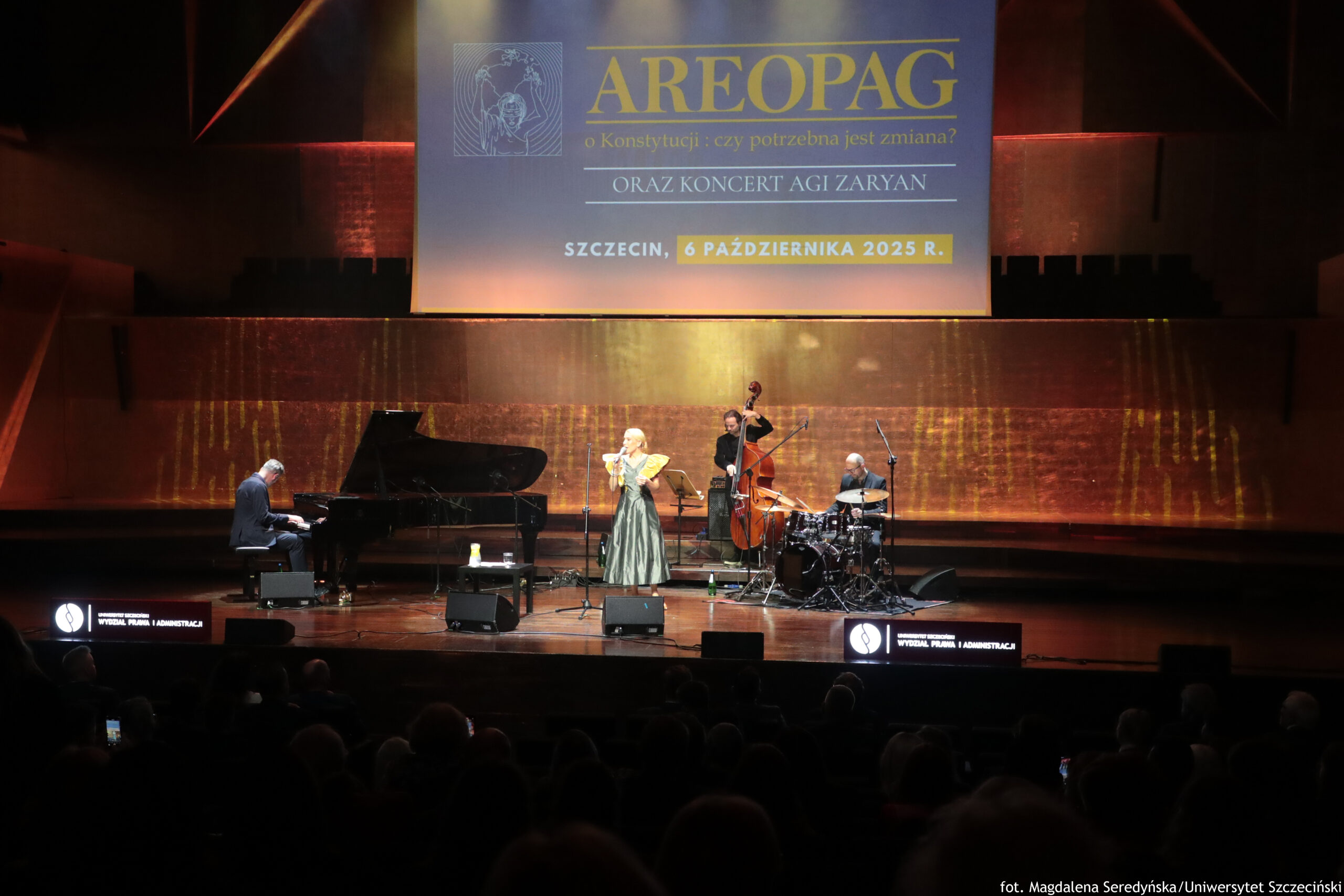 Scena. Występ. Od lewej pianista, wokalistka, kontrabasista i perkusista. Tło złote. Od góry ekran z napisem Areopag o Konstytucji, koncert Agi Zaryan.