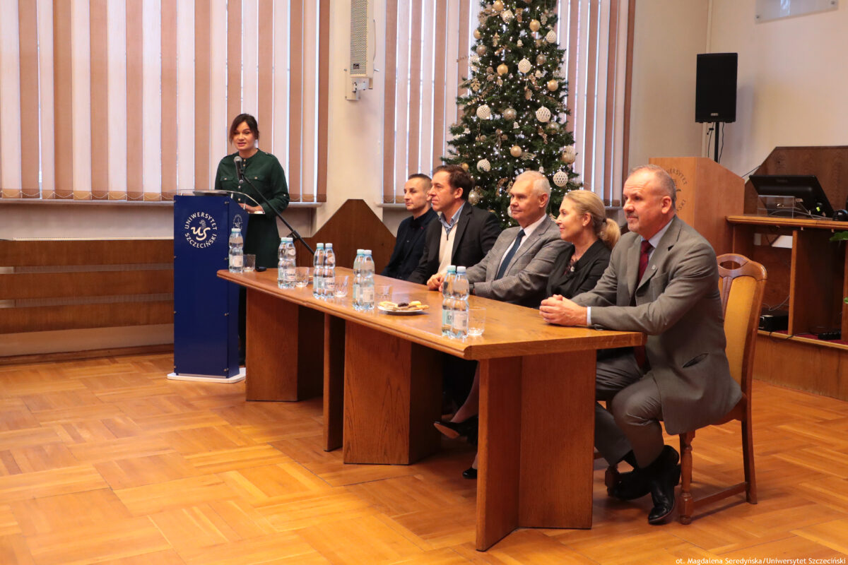 zdjęcie przedstawiające pięcioro osób siedzących przy stole prezydialnym oraz wypowiadającą się przy mównicy kobietę