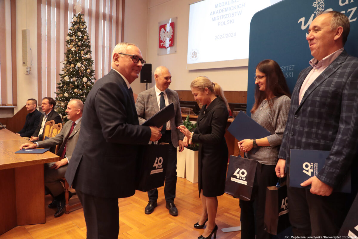 zdjęcie przedstawiające grupę ludzi na pierwszym planie ujęta kobieta oraz mężczyzna podający sobie dłonie i przekazujący teczkę