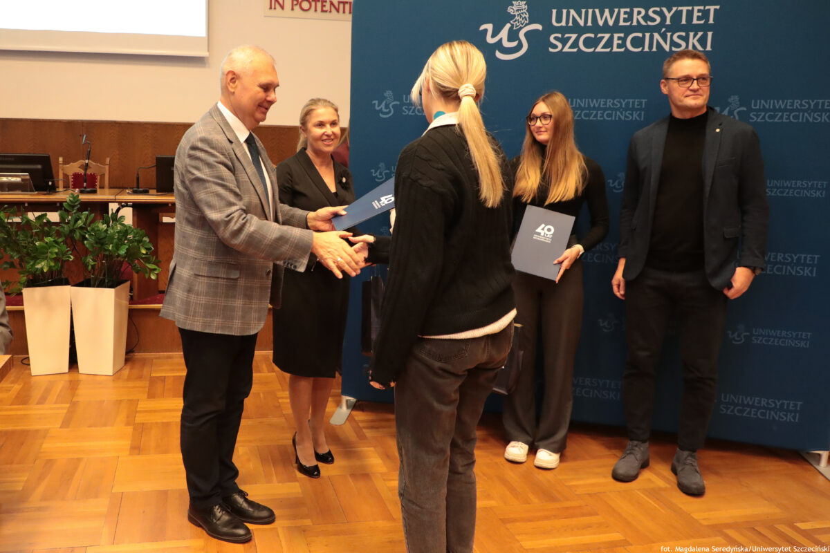 zdjęcie przedstawiające grupę ludzi na pierwszym planie ujęta kobieta oraz mężczyzna podający sobie dłonie i przekazujący teczkę