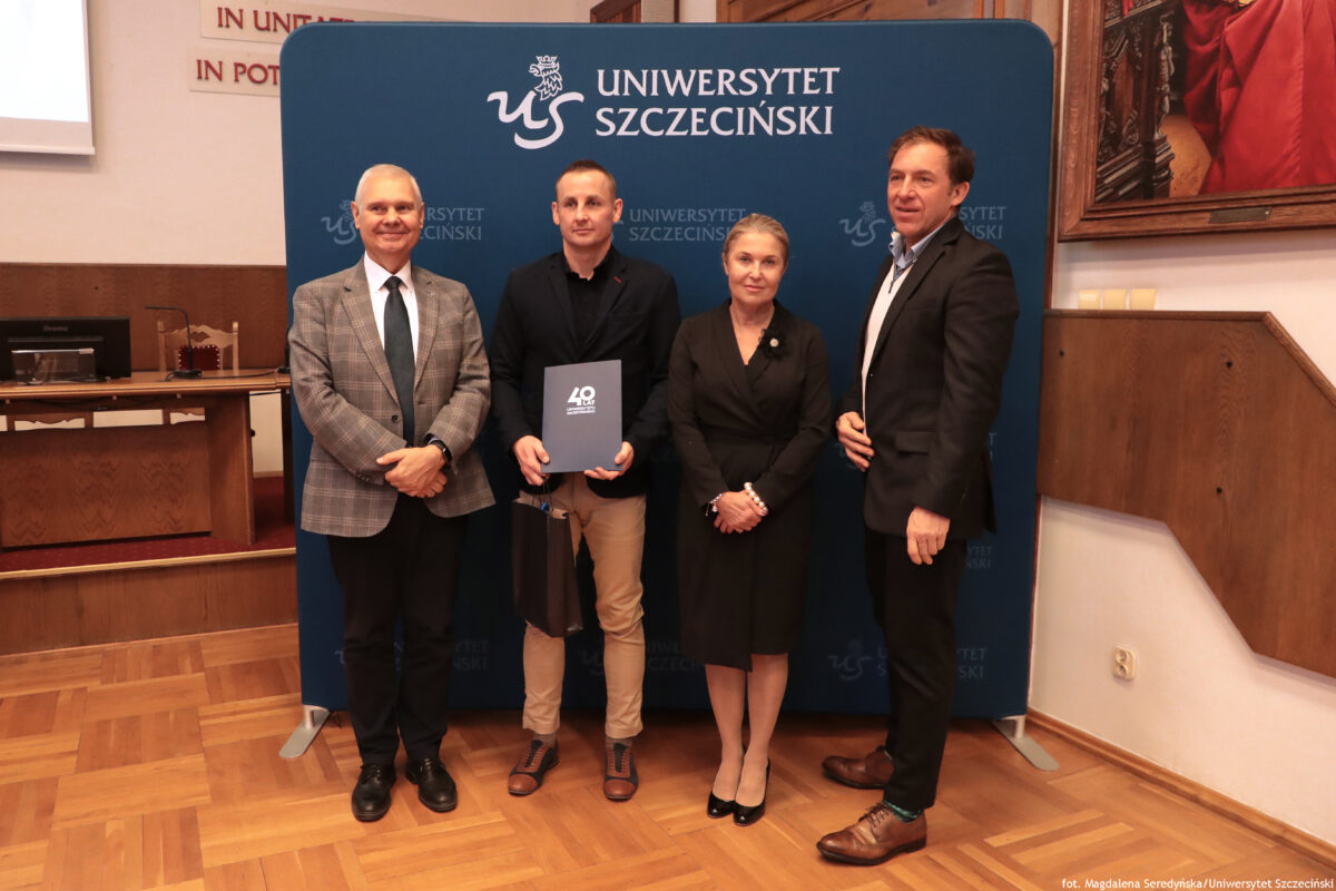 zdjęcie przedstawiające 3 mężczyzn oraz kobietę pozujących na ściance US mężczyzna trzyma w ręku teczkę oraz papierową torbę