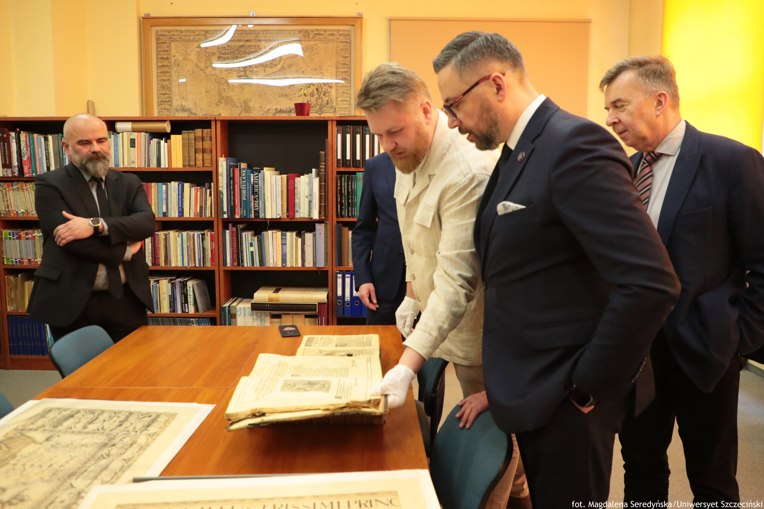 Grupa pięciu mężczyzn w pomieszczeniu przypominającym bibliotekę lub archiwum, stojących przy dużym drewnianym stole z rozłożonymi starymi dokumentami i książką. Na pierwszym planie stoi kilku mężczyzn w garniturach, w średnim wieku; jeden ma krótkie ciemne włosy, brodę i okulary, ubrany jest w ciemny garnitur z białą koszulą i poszetką. Obok niego mężczyzna w jasnej, beżowej koszuli pochyla się nad stołem; na dłoniach ma białe rękawiczki i trzyma otwartą, grubą księgę lub album z pożółkłymi kartami. Z tyłu widać kolejnych mężczyzn w marynarkach; po lewej stoi łysy mężczyzna z gęstą brodą, w ciemnym garniturze, z założonymi rękami. Na stole leży otwarta księga z ilustracjami i tekstem oraz kilka dużych arkuszy papieru (jeden częściowo widoczny na dole kadru). W tle znajdują się regały z książkami i segregatorami, a nad nimi duża oprawiona mapa lub rycina na ścianie. W prawym dolnym rogu zdjęcia jest podpis: „fot. Magdalena Seredyńska/Uniwersytet Szczeciński”.