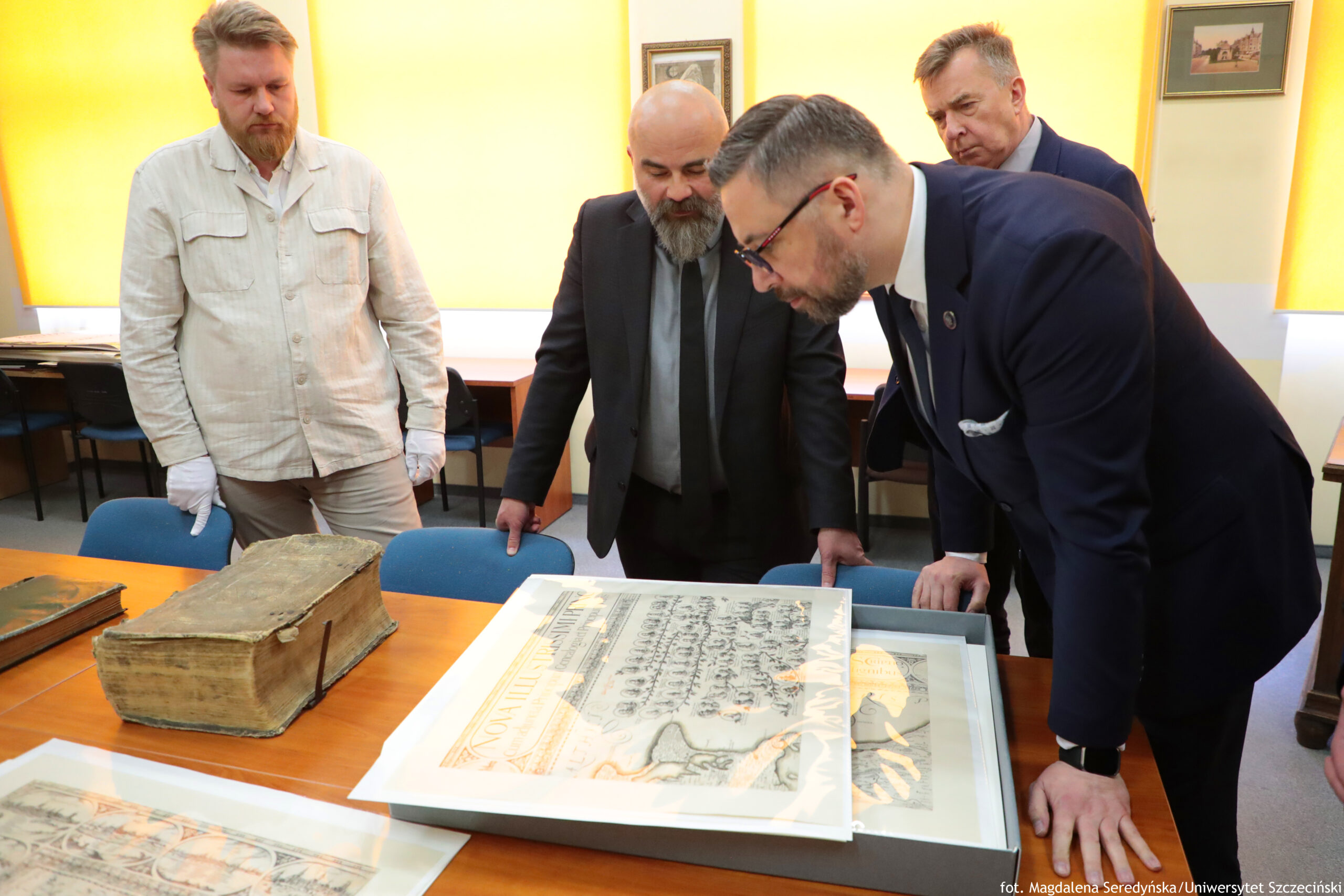 Czterech mężczyzn pochylonych nad dużą, oprawioną kartą lub dokumentem rozłożonym na stole. Pomieszczenie wygląda jak sala biurowa lub konferencyjna, z żółtymi roletami w oknach i krzesłami przy ścianach. Na pierwszym planie po prawej stronie stoi mężczyzna w granatowym garniturze, z krótkimi ciemnymi włosami, okularami i przyciętą brodą; opiera się o stół i przygląda się dokumentowi. Pośrodku stoi mężczyzna łysy z siwą, gęstą brodą, w ciemnym garniturze i krawacie, również patrzący na dokument. Z lewej strony stoi mężczyzna w jasnej, beżowej koszuli i jasnych spodniach, z jasnymi włosami i rudawą brodą; ma na dłoniach białe rękawiczki. Z tyłu, po prawej, stoi kolejny mężczyzna w ciemnym garniturze, z krótkimi włosami, obserwujący to samo. Na stole leży duża mapa lub grafika w ramie albo w płaskiej skrzyni ekspozycyjnej; widać na niej rysunek oraz fragmenty tekstu, ale nie są czytelne w tej rozdzielczości. Po lewej stronie stołu znajduje się duża, stara księga o zniszczonej, pożółkłej okładce i grzbiecie. W dolnym lewym rogu kadru widać jeszcze jeden arkusz lub mapę, częściowo poza kadrem. W prawym dolnym rogu zdjęcia jest mały podpis: „fot. Magdalena Seredyńska/Uniwersytet Szczeciński”.
