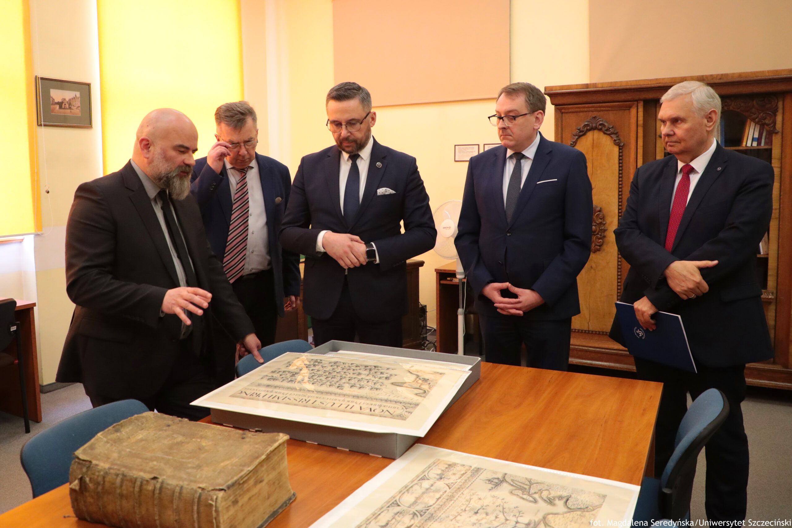 Grupa pięciu mężczyzn w garniturach stojących przy drewnianym stole w pomieszczeniu biurowym, pochylonych nad dużym, otwartym pudełkiem z płaskim dokumentem lub mapą. Po lewej stronie stoi mężczyzna z łysą głową i długą, siwą brodą, w ciemnym garniturze i czarnym krawacie; ma wysuniętą dłoń nad stołem, jakby coś objaśniał. Obok niego stoi mężczyzna w okularach, w garniturze i krawacie w ukośne pasy, z ręką przy twarzy. Na środku stoi mężczyzna z krótką brodą i okularami, w granatowym garniturze, białej koszuli i ciemnym krawacie, z rękami przy mankiecie, jakby sprawdzał zegarek. Dalej stoi mężczyzna w okularach, w ciemnym garniturze i krawacie, z dłońmi złożonymi z przodu. Po prawej stronie stoi mężczyzna o siwych włosach w garniturze z czerwonym krawatem, trzymający granatową teczkę. Na stole są dwa duże płaskie obiekty: w centrum szare pudełko z dużą kartą z nadrukiem (wygląda jak stara mapa lub dokument) oraz niżej na zdjęciu kolejna duża karta z rysunkami. W lewym dolnym rogu leży gruba, stara księga o zniszczonej, brązowej oprawie. W tle widać jasne ściany, obraz w ramce, duży drewniany mebel (szafa lub regał) i mały wiatrak na stojaku. W prawym dolnym rogu jest napis: „fot. Magdalena Seredyńska/Uniwersytet Szczeciński”.