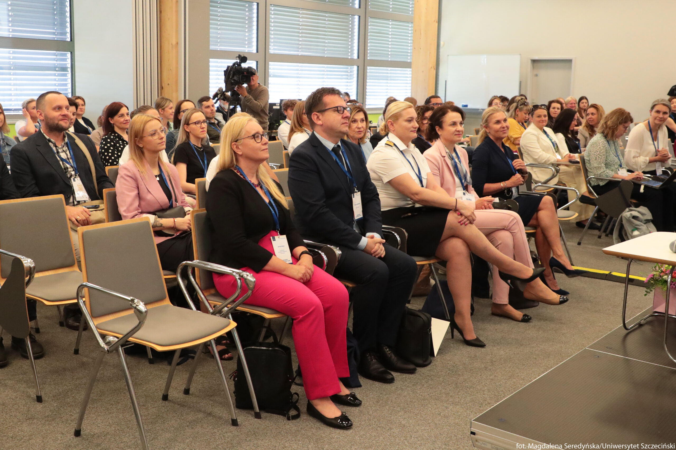Grupa dorosłych osób siedzących w sali konferencyjnej podczas wydarzenia. W pierwszym rzędzie siedzi pięć kobiet i jeden mężczyzna, wszyscy w eleganckich, biznesowych strojach – garnitury, marynarki, koszule, część kobiet w żakietach i garsonkach, jedna ma jaskrawo różowe spodnie. Na szyjach mają niebieskie smycze z identyfikatorami. Krzesła są szare z metalowymi podłokietnikami. Za nimi siedzi kilkadziesiąt kolejnych osób, również w formalnych ubraniach, patrzących w kierunku przodu sali, jakby słuchały prelegenta. Z tyłu po lewej stronie widać kamerzystę z dużą kamerą na statywie, nagrywającego wydarzenie. Tło stanowią duże okna z poziomymi żaluzjami. W prawym dolnym rogu fragment podestu i kawałek stołu. W lewym dolnym rogu, na podłodze przy nogach krzeseł, stoją pojedyncze torby i plecaki. Na brzegu zdjęcia w prawym dolnym rogu jest mały, jasny napis z informacją o autorce zdjęcia i uczelni.