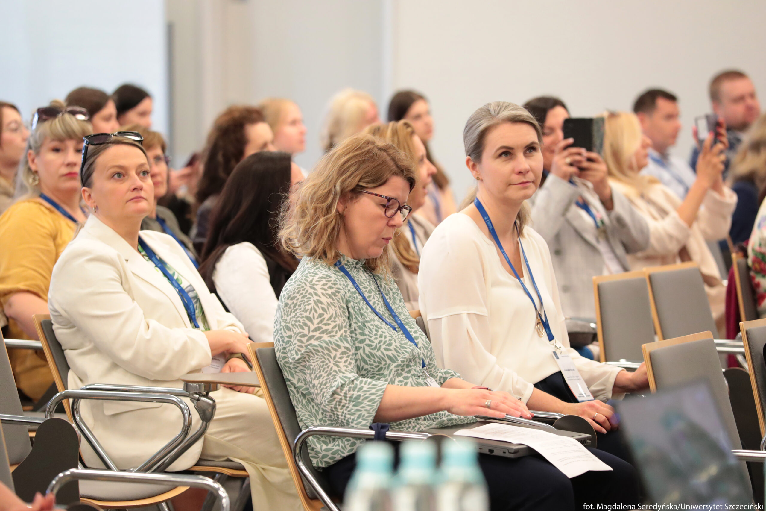 Grupa osób siedzących na sali konferencyjnej. Na pierwszym planie, z lewej strony, siedzi kobieta w białym garniturze i niebieskim topie, z jasną cerą i włosami związanymi do góry; na głowie ma okulary przeciwsłoneczne, na szyi niebieską smycz z identyfikatorem. Obok niej, bliżej środka, siedzi kobieta o jasnej cerze i falowanych, jasnobrązowych włosach do ramion; ma okulary i bluzkę w jasnozielony roślinny wzór, w rękach trzyma kartki z notatkami leżące częściowo na kolanach. Po prawej stronie pierwszego planu znajduje się kobieta o jasnej cerze i prostych, siwiejących włosach związanych w niski kucyk; ma na sobie jasną, kremową bluzkę z długimi rękawami i również smycz z identyfikatorem. W drugim i trzecim rzędzie siedzi wiele innych osób, głównie kobiety, ubrane elegancko lub półformalnie; część z nich trzyma telefony, część patrzy przed siebie, inne spoglądają w dół. Krzesła mają metalowe podłokietniki i szare oparcia. Na dole kadru, na pierwszym planie, widać rozmyte butelki wody oraz fragment otwartego laptopa.
