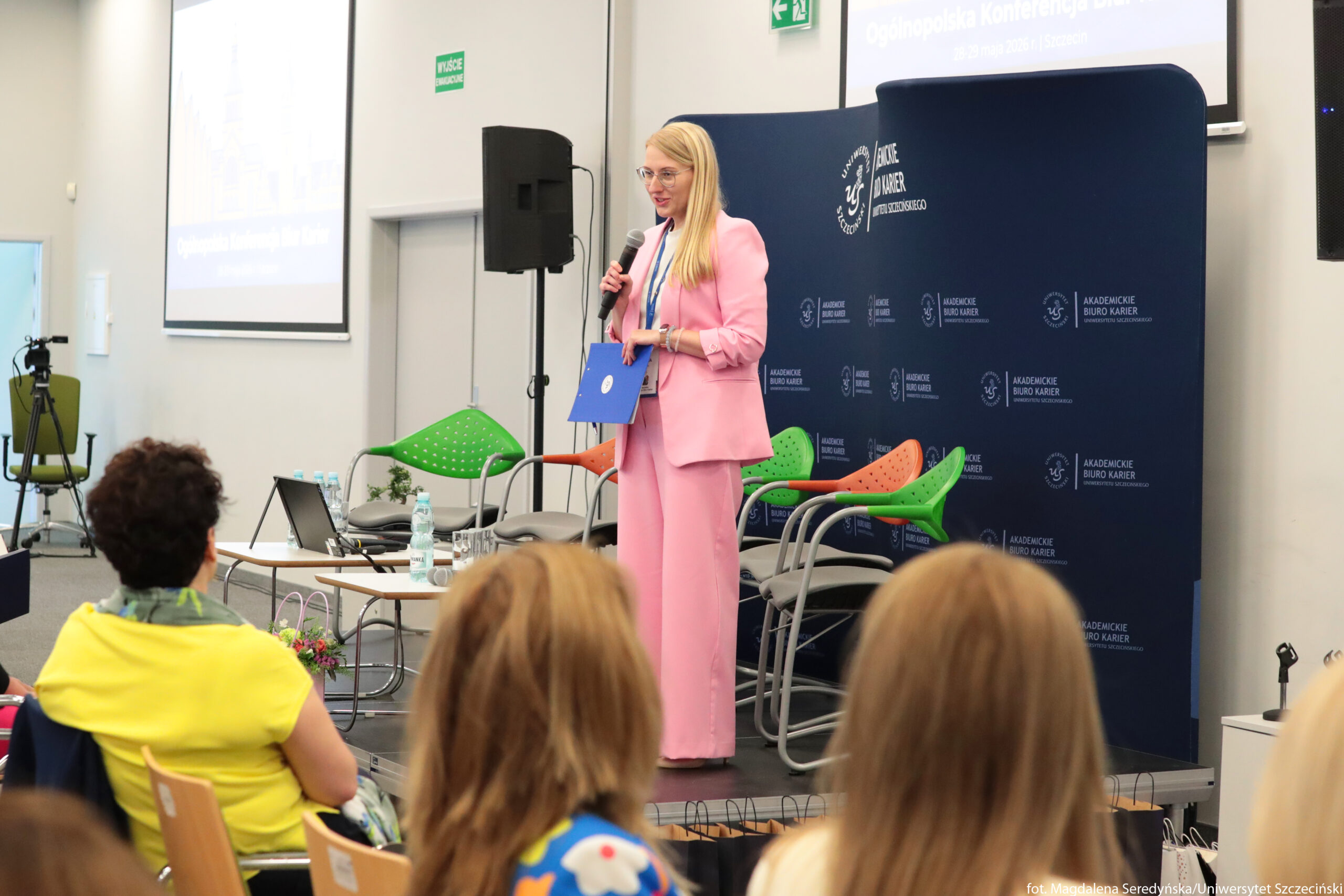 Kobieta w różowym garniturze stojąca na scenie z mikrofonem w dłoni. Ma długie blond włosy, jasne okulary i trzyma niebieską teczkę lub segregator. Stoi przed ciemnoniebieską ścianką z białymi logotypami „Akademickie Biuro Karier” i herbem Uniwersytetu Szczecińskiego. W tle znajduje się wiszący ekran projekcyjny z prezentacją, której tekst w górnej części jest słabo czytelny, widać jednak napis „Ogólnopolska Konferencja…”. Na scenie stoją kolorowe krzesła – zielone i pomarańczowe z oparciami w kropki oraz szare krzesła biurowe. Przed sceną, na pierwszym planie, siedzą tyłem do obiektywu uczestnicy wydarzenia, widoczne są głowy i ramiona kilku osób. Na stoliku po lewej stronie sceny leżą plastikowe butelki wody, szklanki i otwarty laptop. Po lewej stronie sali stoi statyw z kamerą skierowaną w stronę sceny. Na ścianie za sceną wisi zielony znak „Wyjście ewakuacyjne”.