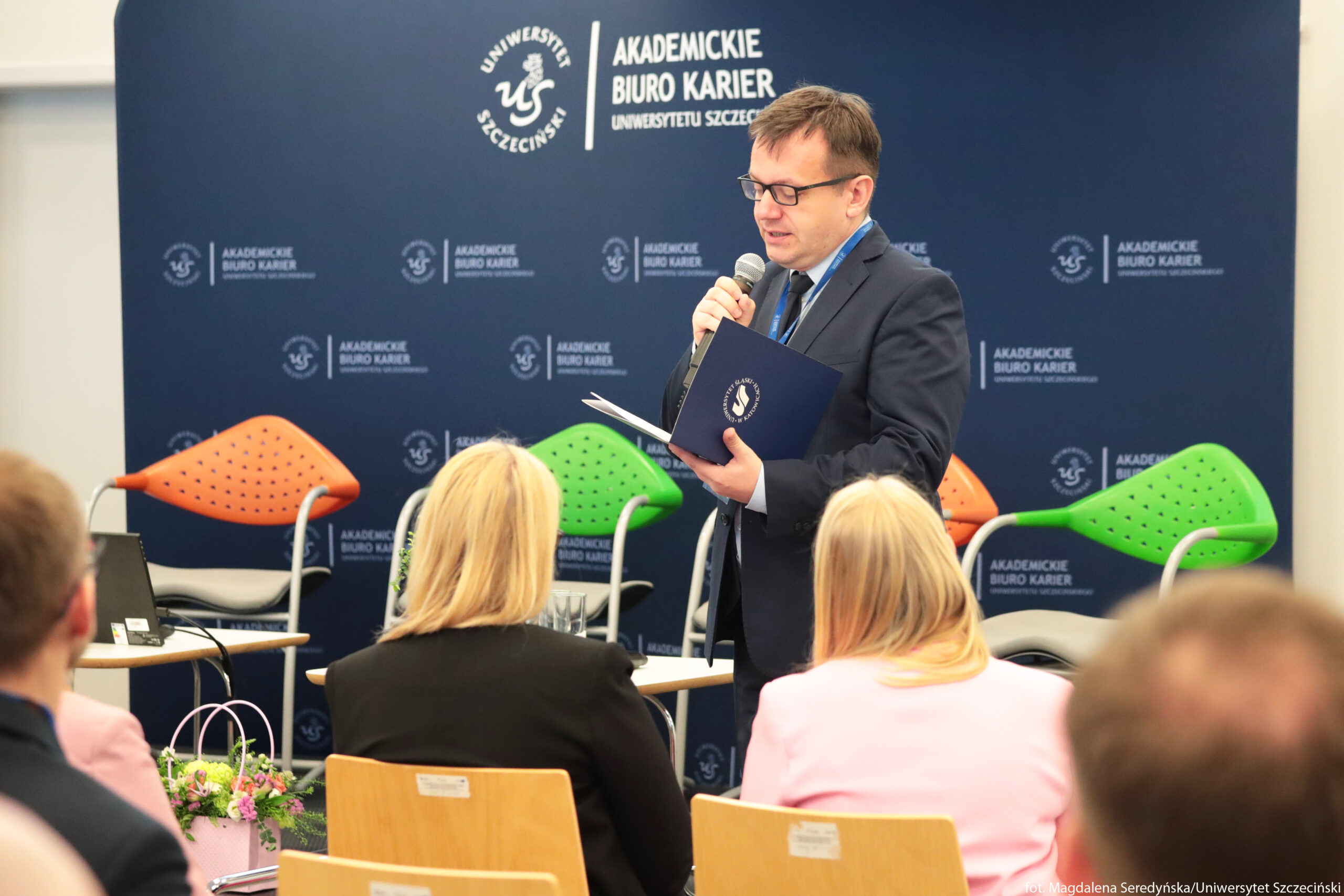 Mężczyzna w garniturze przemawiający do publiczności z mikrofonem w dłoni. Stoi z prawej strony zdjęcia, trzyma otwartą granatową teczkę z logo Uniwersytetu Szczecińskiego, z której najwyraźniej czyta. Ma krótkie brązowe włosy, okulary i jasną skórę. Za nim znajduje się granatowa ścianka z białymi napisami i logotypami: „UNIWERSYTET SZCZECIŃSKI” oraz „AKADEMICKIE BIURO KARIER UNIWERSYTETU SZCZECIŃSKIEGO”. Napis i logo powtarzają się w kilku miejscach tła. Na scenie stoją kolorowe krzesła konferencyjne o ażurowych oparciach: po lewej i prawej stronie widoczne są krzesła w kolorze pomarańczowym i zielonym. Przed sceną siedzi publiczność – z tyłu widać trzy osoby z jasnymi włosami i jasną skórą, ubrane w marynarki w kolorze czarnym i jasnoróżowym. Przed pierwszym rzędem krzeseł, po lewej stronie, stoi mały stolik z otwartym laptopem i przewodami. Na podłodze obok znajduje się różowa torebka prezentowa wypełniona kwiatami w pastelowych kolorach. W prawym dolnym rogu widoczny jest biały, mały napis z informacją o autorce zdjęcia i uczelni.