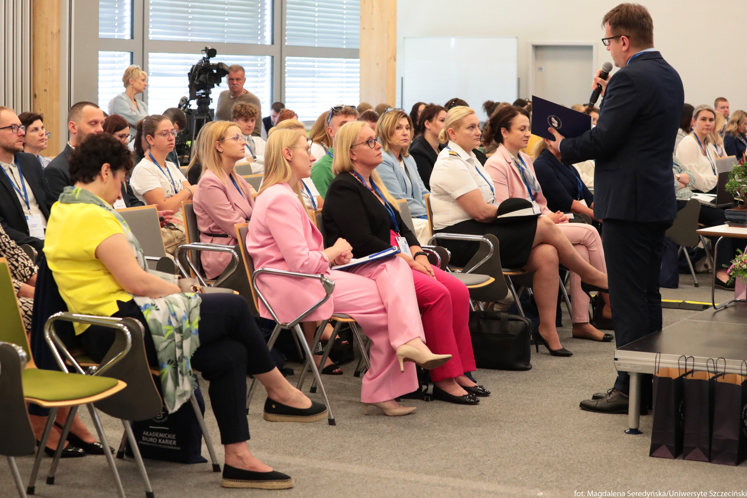 Sala konferencyjna wypełniona siedzącymi osobami i mężczyzna mówiący do mikrofonu z przodu. Publiczność siedzi w kilku rzędach na krzesłach z metalowymi podłokietnikami, większość osób ma identyfikatory na niebieskich smyczach. Na pierwszym planie widać głównie kobiety w formalnych strojach – między innymi jasnoróżowy garnitur, czarne spodnie z żółtą bluzką i szal w kwiatowy wzór, jasnoróżową marynarkę, czarną marynarkę z różowymi spodniami oraz białą koszulę mundurową z czarną spódnicą. Po prawej stronie, na podwyższeniu, stoi mężczyzna w granatowym garniturze, trzyma mikrofon jedną ręką, a drugą niebieską teczkę lub skoroszyt, z którego czyta. Osoby na widowni patrzą w jego stronę, część trzyma notatniki lub teczki na kolanach. W tle, przy dużych oknach z poziomymi żaluzjami, stoi kamera na statywie obsługiwana przez jednego mężczyznę, obok niego stoi kobieta w jasnym stroju. Podłoga jest wykładzina w szarym kolorze, a po prawej przy scenie stoją ciemne papierowe torby prezentowe.