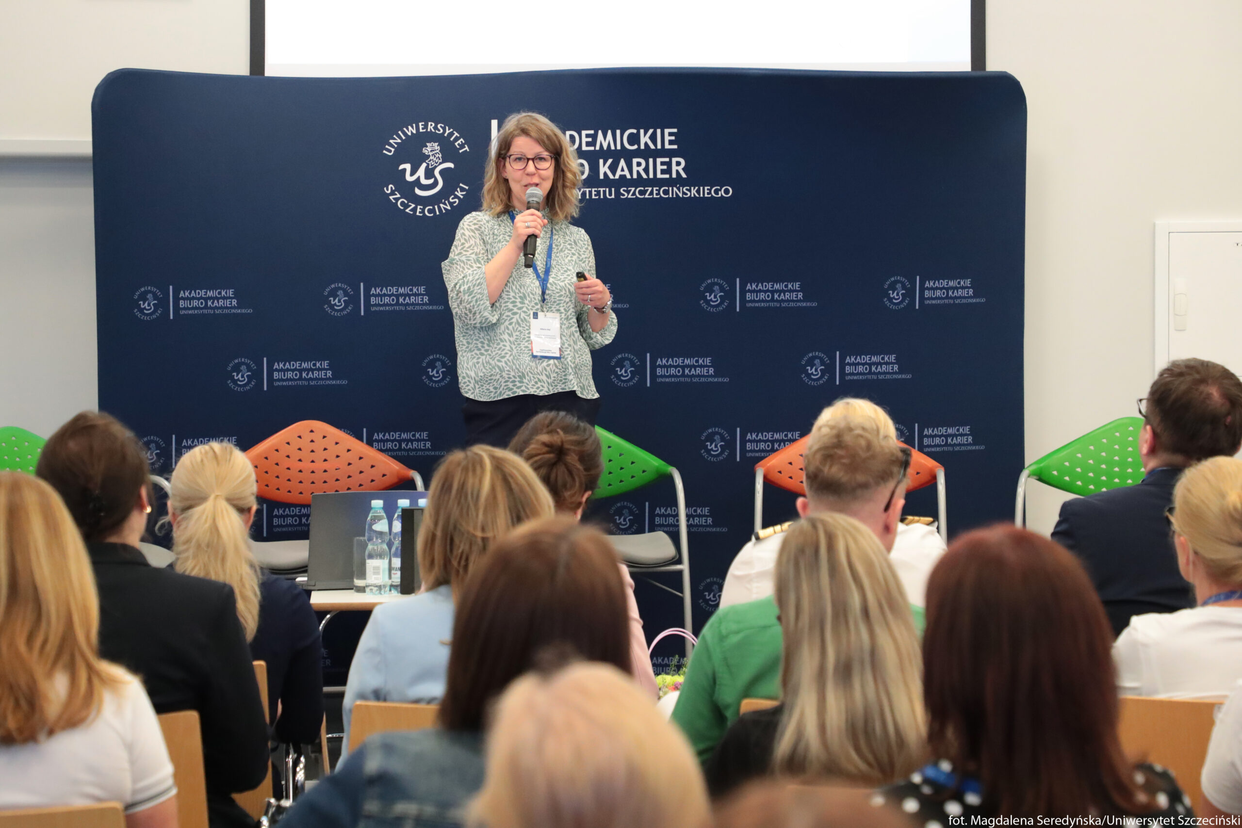 Kobieta stojąca na scenie przed publicznością. Na środku zdjęcia stoi kobieta o jasnej skórze i średniej długości falowanych, jasnobrązowych włosach, w okularach. Ubrana jest w luźną, wzorzystą, jasnozielono‑niebieską bluzkę i ciemne spodnie. W prawej dłoni trzyma mikrofon, a w lewej niewielki przedmiot, prawdopodobnie pilot lub wskaźnik. Na szyi ma niebieską smycz z identyfikatorem. Za nią znajduje się duża, ciemnoniebieska ścianka z białymi napisami i logotypami Uniwersytetu Szczecińskiego oraz napisem „Akademickie Biuro Karier”. Przed ścianką stoją kolorowe krzesła – po lewej pomarańczowe, pośrodku zielone, po prawej kolejne pomarańczowe i zielone. Na pierwszym planie widać tyły głów kilkunastu osób siedzących na krzesłach, zwróconych w stronę prelegentki; mają różne kolory włosów, większość w odcieniach blond i brązu. Na stole z przodu stoją plastikowe butelki z wodą i szklanki. W prawym dolnym rogu znajduje się mały biały napis: „fot. Magdalena Seredyńska/Uniwersytet Szczeciński”.