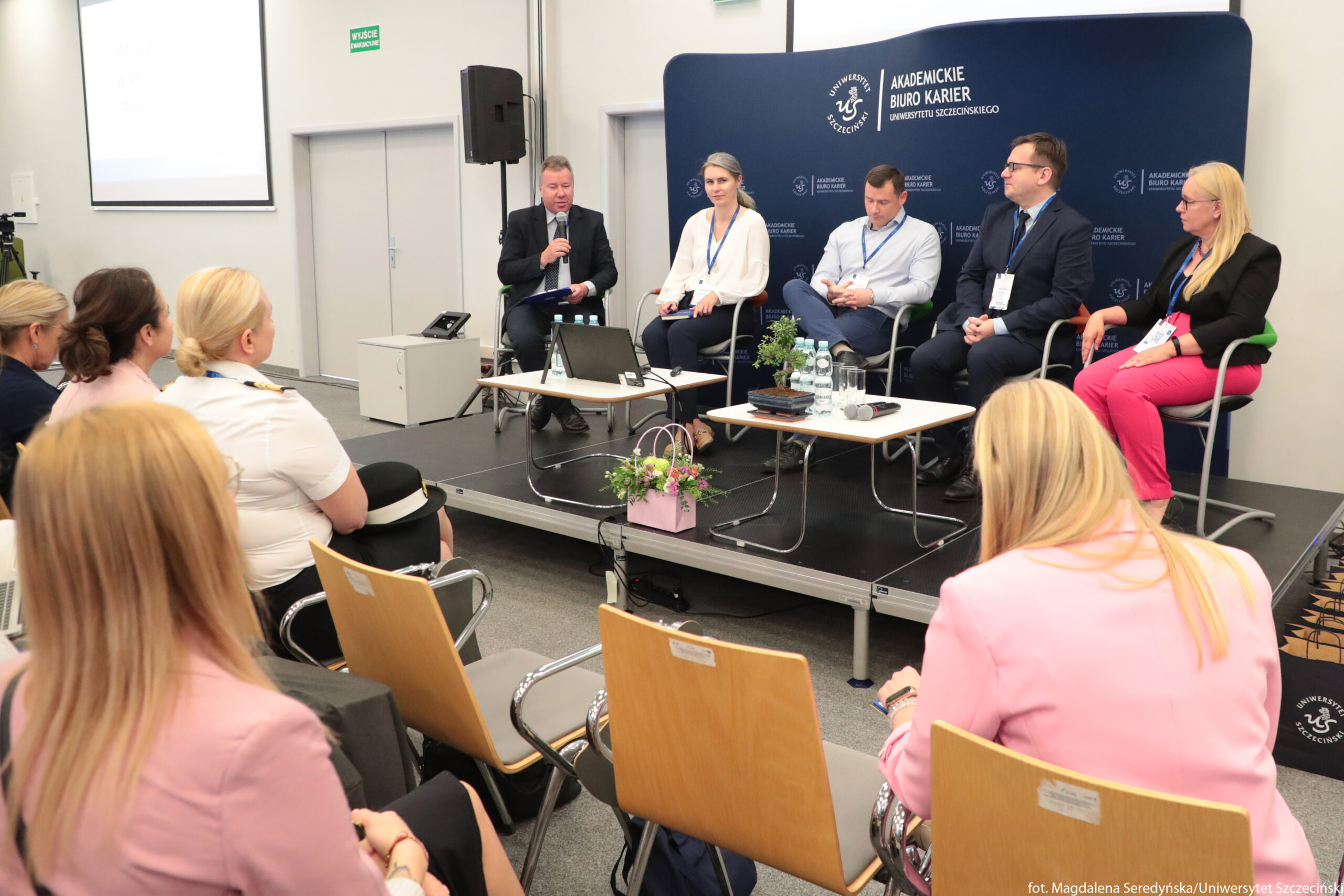 Panel dyskusyjny na konferencji w sali uczelni. Na podwyższonej scenie siedzi pięć osób przy prostokątnych stolikach; czterech mężczyzn i jedna kobieta, wszyscy w biznesowych lub półformalnych strojach z identyfikatorami na niebieskich smyczach. Po lewej stronie panelu mężczyzna w garniturze trzyma mikrofon i mówi, obok niego siedzi kobieta w białej bluzce, dalej dwóch mężczyzn w jasnych koszulach i granatowych garniturach, a z prawej kobieta w czarnej marynarce i różowych spodniach. Na stolikach stoją butelki wody, szklanki, teczki i laptop, a między stolikami stoi różowa papierowa torebka z bukietem kwiatów. Za panelistami znajduje się ciemnoniebieska ścianka z logo i napisem „Akademickie Biuro Karier Uniwersytetu Szczecińskiego” oraz symbolem uczelni. Przed sceną siedzi publiczność na drewnianych krzesłach z metalowymi podłokietnikami; widać kilka kobiet w jasnych marynarkach i jedną osobę w białym mundurze z krótkim rękawem. Po lewej stronie kadru znajduje się kamera na statywie, a z tyłu ściana z zamkniętymi, podwójnymi drzwiami i zielonym napisem „WYJŚCIE”.