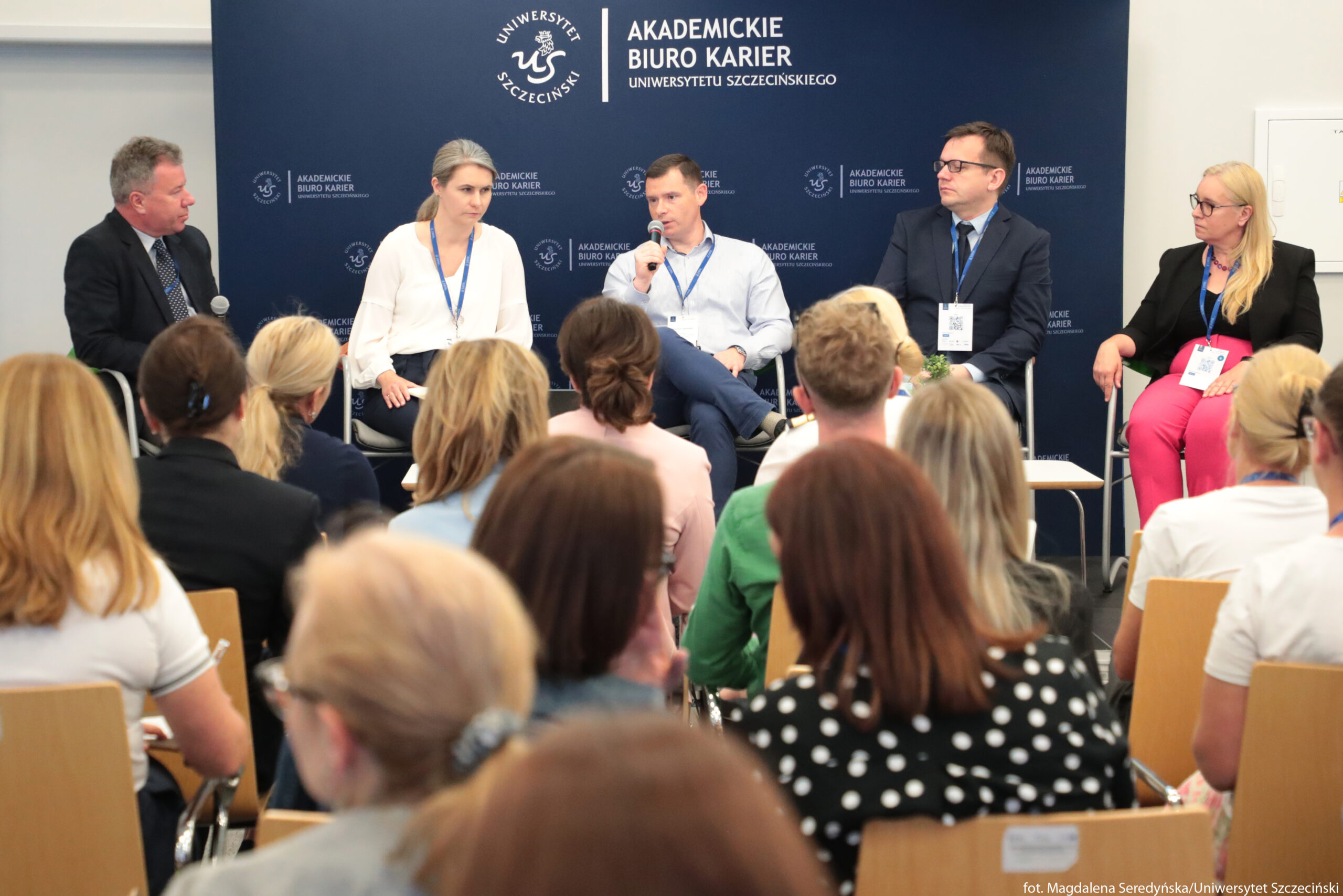 Panel dyskusyjny z pięcioma prelegentami siedzącymi na scenie przed publicznością. Na pierwszym planie znajdują się rzędy osób siedzących na drewnianych krzesłach, widocznych głównie od tyłu; większość ma jasne lub średnie włosy, część spiętych, część rozpuszczonych. Na scenie, na tle granatowej ścianki, siedzi pięć osób. Po lewej stronie siedzi mężczyzna o jasnej skórze, siwych włosach, w ciemnym garniturze i koszuli, trzymający mikrofon. Obok niego siedzi kobieta o jasnej skórze i jasnych, upiętych włosach, w białej bluzce i ciemnych spodniach, z niebieską smyczą na szyi. Na środku siedzi mężczyzna o jasnej skórze, w błękitnej koszuli i ciemnych spodniach, trzymający mikrofon przy ustach, jakby właśnie mówił. Dalej po prawej siedzi mężczyzna o jasnej skórze, w ciemnym garniturze, jasnej koszuli i okularach, z identyfikatorem na smyczy. Najbardziej po prawej siedzi kobieta o jasnej skórze i długich blond włosach, w czarnej marynarce i różowych spodniach, także z identyfikatorem na smyczy.