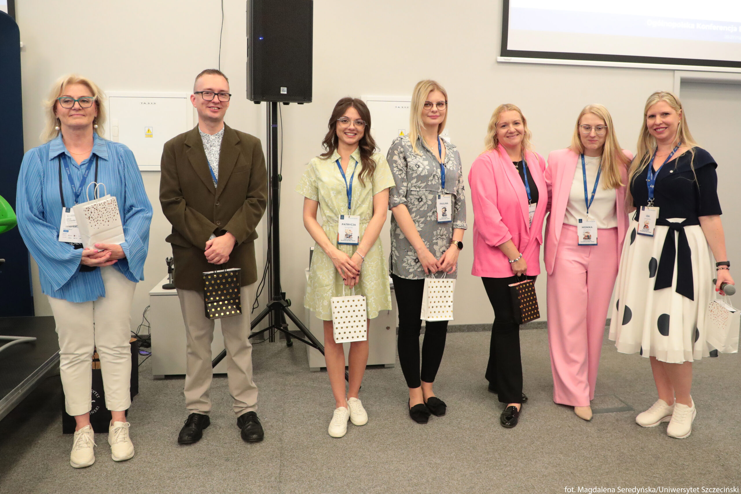 Grupa siedmiu dorosłych osób stojących obok siebie w jasnym pomieszczeniu konferencyjnym. Wszyscy ustawieni są w szeregu przodem, mają identyfikatory na niebieskich smyczach i w dłoniach trzymają ozdobne torebki prezentowe – białe lub czarne z drobnym wzorem. Od lewej: kobieta o jasnych włosach do ramion, w okularach, ubrana w luźną niebieską koszulę z długimi rękawami, jasne spodnie i białe sportowe buty. Obok stoi mężczyzna o krótkich włosach, w okularach, w brązowej marynarce, jasnych spodniach i czarnych butach; ma jasnoszarą koszulę z drobnym wzorem. Trzecia osoba to młoda kobieta o średniej długości brązowych włosach, w jasnej, krótkiej sukience z delikatnym wzorem i białych tenisówkach. Czwarta kobieta ma proste blond włosy do ramion, nosi szarą tunikę z roślinnym wzorem, czarne dopasowane spodnie i czarne buty. Piąta kobieta ma dłuższe falowane blond włosy, ma na sobie luźną różową koszulę, czarne spodnie i czarne półbuty. Szósta kobieta ma długie proste blond włosy, ubrana jest w jasnoróżowy garnitur – marynarkę i spodnie – oraz białą bluzkę i beżowe buty na niskim obcasie; stoi z ręką opartą na ramieniu sąsiadki. Siódma kobieta ma długie blond włosy, granatową górę z krótkim rękawem i rozkloszowaną białą spódnicę w duże granatowe grochy, przewiązaną granatowym paskiem; na stopach ma białe sportowe buty i trzyma mikrofon oraz białą torebkę prezentową. W tle widać fragment ekranu projekcyjnego oraz głośnik na stojaku. Całość sprawia wrażenie uroczystego zakończenia lub wyróżnienia uczestników konferencji.