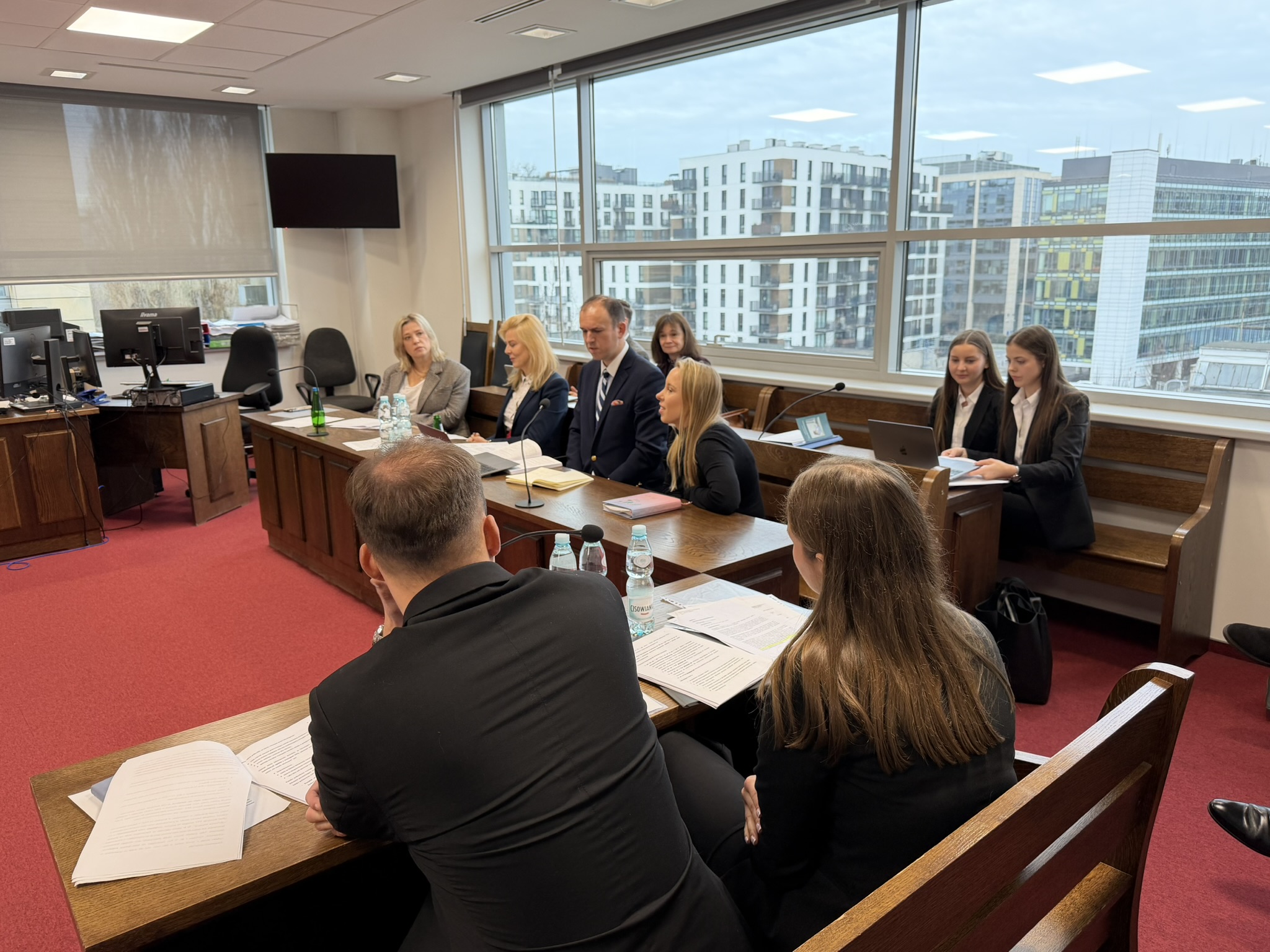 Pomieszczenie do symulacji rozprawy sądowej. Za drewnianymi zabudowanymi stołami siedzi głównie młodzież – przed niektórymi stoją laptopy lub tablety.