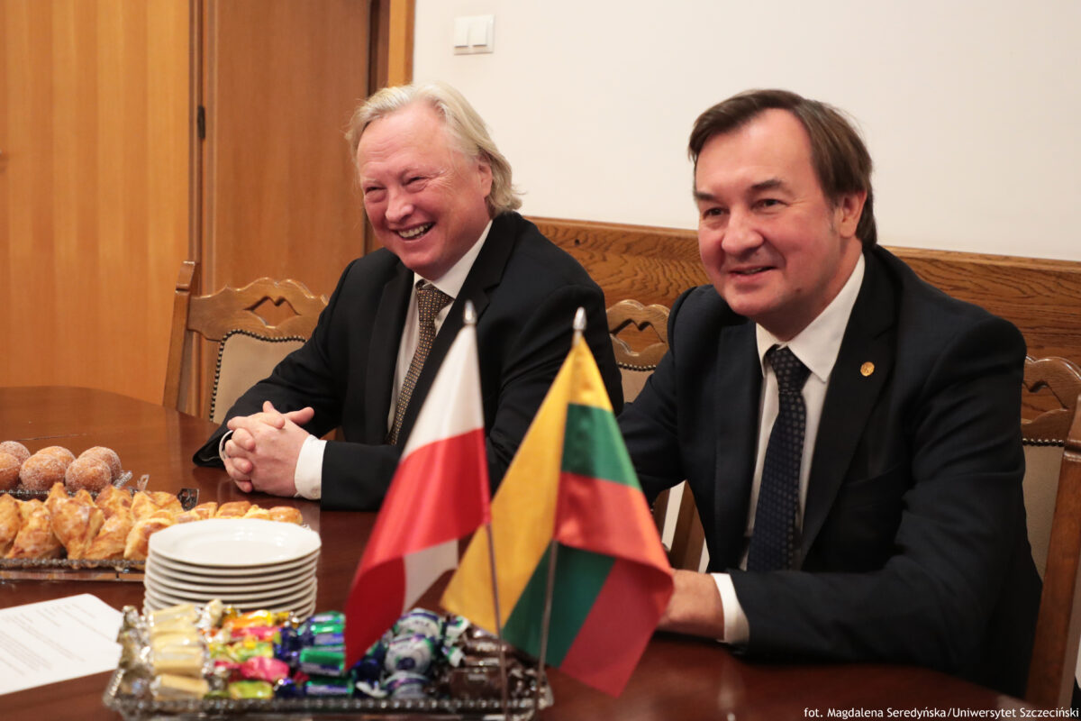 Rektor i prorektor Uniwersytetu Wileńskiego siedzą przy stole, na którym stoją flagi – litewska i polska. Obok cukierki, ciastka i talerzyki.