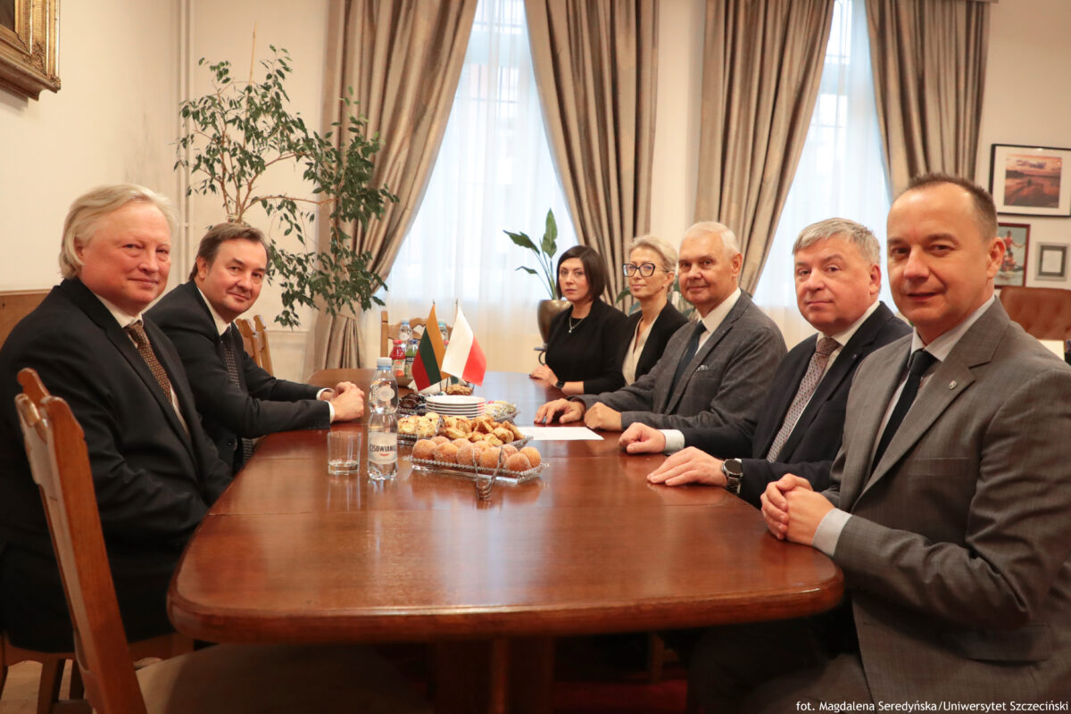 Ujęcie bokiem przy stole. Po lewej dwóch przedstawicieli Uniwersytetu Wileńskiego, po prawej pięcioro przedstawicieli Uniwersytetu Szczecińskiego (dwie kobiety, trzech mężczyzn – w tym rektor i prorektor). Na stole flagi obu krajów i ciastka.