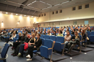 Ujęcie bokiem. Aula. Na niebieskich fotelach siedzi kilkudziesięcioosobowa grupa studentów – uczestników Dni Adaptacyjnych.