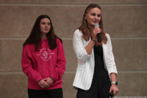 Aula. Dwie przedstawicielki uczelni (ujęcie do połowy) – z przodu kobieta z mikrofonem, z tyłu kobieta w różowej bluzie z białym okrągłym logiem Uniwersytetu Szczecińskiego.