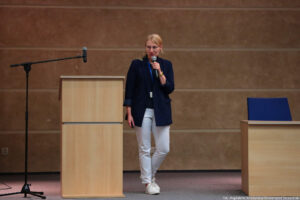 Aula. Po lewej mównica, po prawej biurko z krzesłem. Na środku kierowniczka Akademickiego Biura Karier – Monika Smoguła – trzymająca mikrofon i przybliżająca działanie Biura uczestnikom Dni Adaptacyjnych.