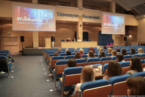 Aula (ujęcie bokiem). Tyłem siedzący na niebieskich fotelach studenci. W tle podest z mównicą i stołem, za którym siedzi troje przedstawicieli uczelni. Na podeście obok mównicy kierowniczka Akademickiego Biura Karier – Monika Smoguła – trzymająca mikrofon i przybliżająca działanie Biura uczestnikom Dni Adaptacyjnych. Od góry niebieski napis: Uniwersytet Szczeciński, po bokach dwa ekrany z napisem #FunRunUS, 22 października 2025 r.