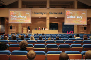 Aula. Tyłem siedzący na niebieskich fotelach studenci. W tle podest z mównicą i stołem, za którym siedzi troje przedstawicieli uczelni. Na podeście obok mównicy kierowniczka Akademickiego Biura Karier – Monika Smoguła – trzymająca mikrofon i przybliżająca działanie Biura uczestnikom Dni Adaptacyjnych. Od góry niebieski napis: Uniwersytet Szczeciński, po bokach dwa ekrany z napisem Giełda Pracy, 18 listopada 2025 r.