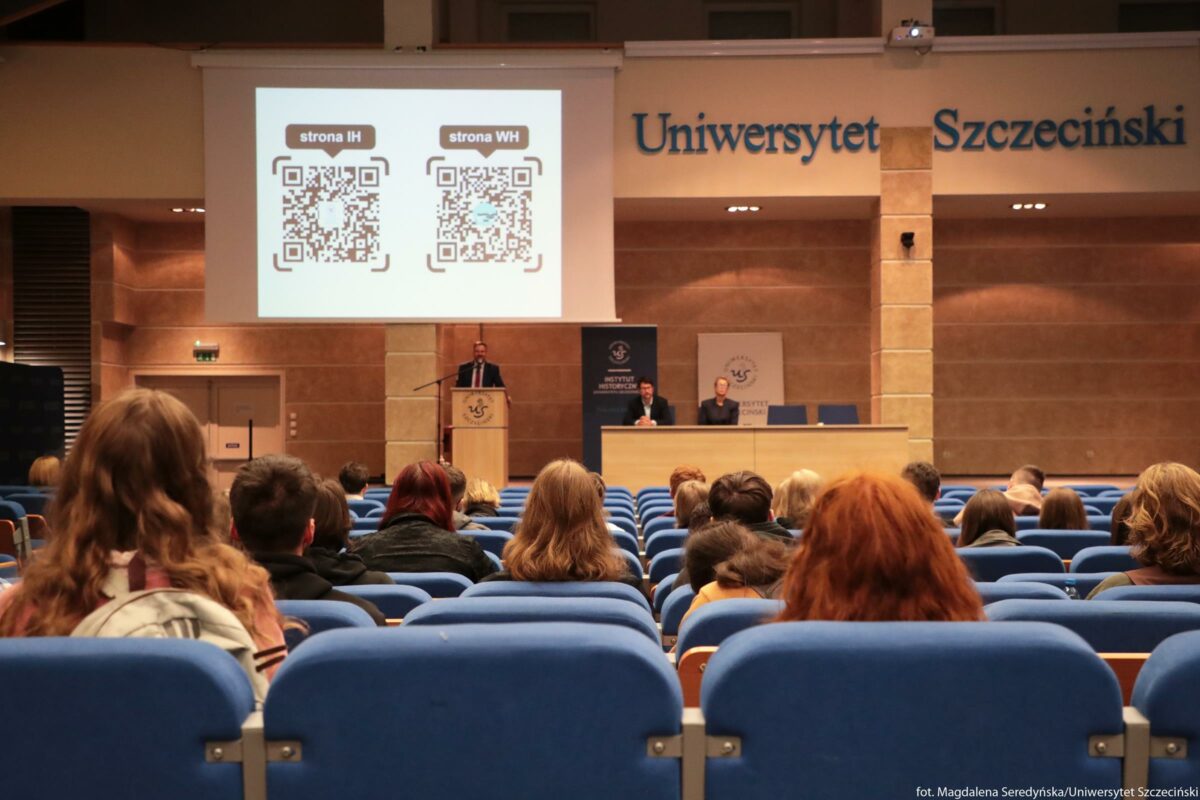 Sala wykładowa Uniwersytetu Szczecińskiego z publicznością siedzącą w niebieskich fotelach, skierowaną w stronę podium i ekranu. Na jasnym ekranie projekcyjnym widoczne są dwa duże kody QR obok siebie. Nad lewym kodem QR jest napis „strona IH”, nad prawym „strona WH”. Pod ekranem stoi mówca przy mównicy ustawionej centralnie; na mównicy widnieje logo Uniwersytetu Szczecińskiego. Po prawej stronie, za długim stołem, siedzą dwie osoby; za nimi stoją dwa pionowe banery z logo i napisem „Instytut Historyczny” (tekst na środkowym banerze jest dobrze widoczny, na prawym częściowo). Na ścianie nad sceną duży napis z niebieskich liter: „Uniwersytet Szczeciński”. W kadrze widocznych jest kilkadziesiąt osób od tyłu, w różnym wieku, większość ma na sobie jesienne lub zimowe ubrania w stonowanych kolorach.