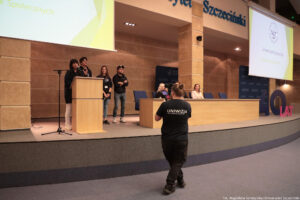 Aula. Podest z mównicą i stołem, za którym siedzi troje przedstawicieli uczelni. Za mównicą Beata Mikołajewska-Wieczorek – dyrektorka Centrum Edukacji Medialnej i Interaktywności. Za nią troje przedstawicieli jednostki. Na pierwszym planie kamerzysta (tyłem do kadru) z białym napisem Uniwizja na koszulce.