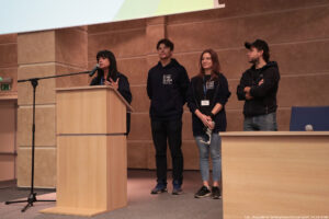 Aula. Podest z mównicą, za którą stoi Beata Mikołajewska-Wieczorek – dyrektorka Centrum Edukacji Medialnej i Interaktywności. Za nią (po prawej) troje przedstawicieli jednostki (dwóch chłopaków i dziewczyna w środku) z napisami Uniwizja na bluzach i koszulkach.