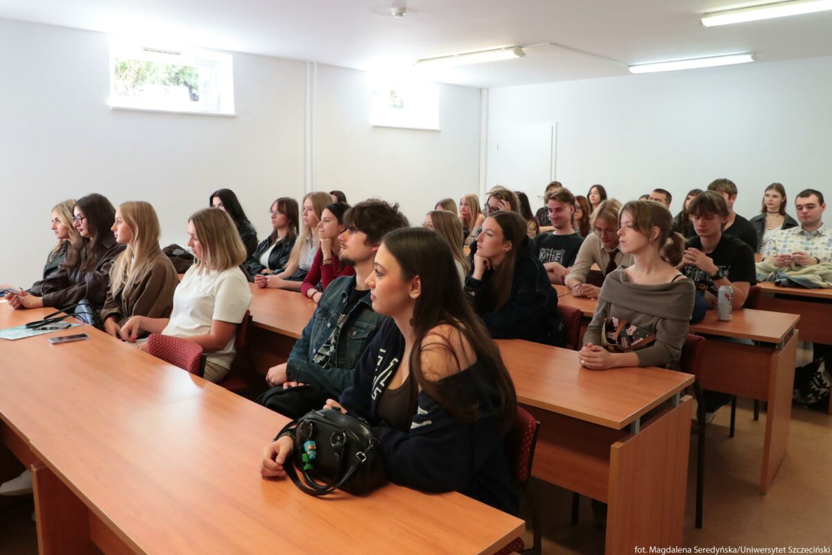Sala wykładowa z kilkoma rzędami drewnianych ławek, przy których siedzi grupa młodych dorosłych. W sali jest jasno, z białymi ścianami i prostokątnymi oknami wysoko pod sufitem, przez które wpada dzienne światło, a na suficie świecą długie jarzeniówki. Przy ławkach siedzi kilkadziesiąt osób, głównie młode kobiety i mężczyźni o jasnej i śniadej karnacji, w swobodnych, codziennych ubraniach: dżinsowych kurtkach, bluzach, swetrach, t‑shirtach. Wszyscy skierowani są w jedną stronę, w stronę niewidocznego prowadzącego, mają poważne lub skupione miny, część opiera brodę na dłoni, inni mają ręce splecione na blacie. Na stołach leżą pojedyncze przedmioty: telefony, zeszyty, kubek termiczny, a przy pierwszej ławce widać czarną torebkę z przypiętymi kolorowymi brelokami. Tło jest proste i puste, bez dekoracji, co podkreśla typowy, uniwersytecki charakter pomieszczenia.