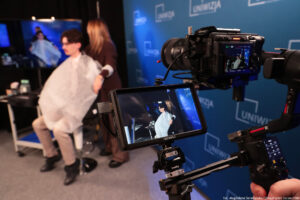 Po prawej kamera z ujęciem na małym ekranie. W tle filmowany obraz – młody mężczyzna siedzący bokiem na hokerze w założonej jasnej pelerynie i kobieta rozkładająca pelerynę. W tle kamery granatowa ścianka z niebieskimi logami Uniwizji.