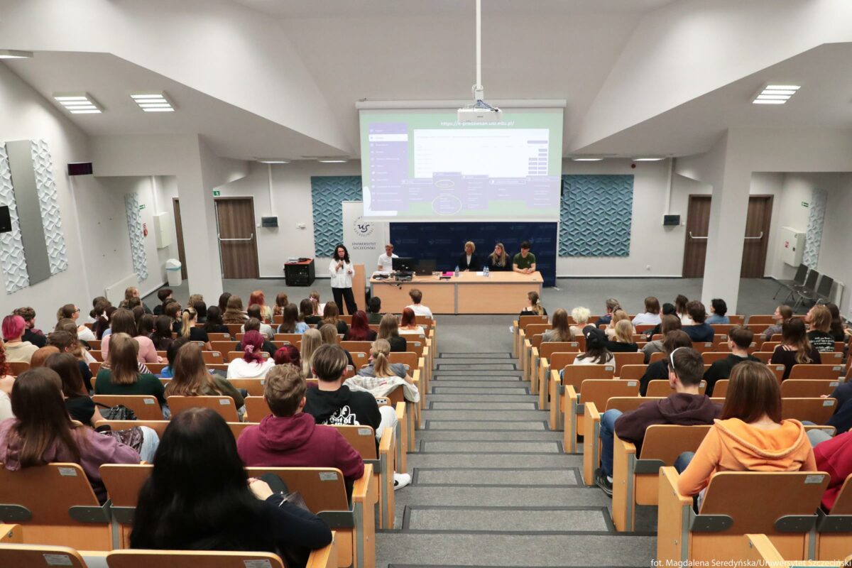 Duża sala wykładowa z pełnymi rzędami studentów siedzących na drewnianych krzesłach w amfiteatralnym układzie. Studenci zwróceni są w stronę przodu sali, gdzie stoi prowadząca w białej koszuli, przemawiająca przed tablicą i ekranem projekcyjnym. Przed ekranem ustawiony jest długi stół prezydialny z czterema osobami siedzącymi za nim; na stole leży laptop, butelki wody i dokumenty. Nad stołem wyświetlana jest prezentacja w odcieniach zieleni i fioletu; na górze widać adres strony internetowej „https://do.gumieszkam.usz.edu.pl/”. Po lewej stronie ekranu stoi biały roll‑up z logotypem Uniwersytetu Szczecińskiego i napisem „UNIWERSYTET SZCZECIŃSKI”. Ściana za stołem ma dekoracyjne panele w niebieskawym kolorze. Po bokach sali znajdują się drzwi z drewnianymi skrzydłami, lampy sufitowe i elementy akustyczne na ścianach. Na schodach pośrodku sali biegnie przejście w dół, a po obu jego stronach zajęte są wszystkie miejsca przez uczestników wykładu lub spotkania.