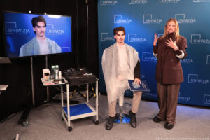 Na środku młody mężczyzna siedzący na hokerze w założonej jasnej pelerynie. Po prawej kobieta opowiadająca o przygotowaniu osób do występu na wizji. Po lewej kwadratowy, piętrowy stolik na kółkach, na górze którego leżą niezbędne akcesoria. W tle granatowa ścianka z niebieskimi logami Uniwizji. Po lewej duży ekran, na którym widać wspomnianego mężczyznę.