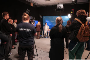 Na pierwszym planie stojące tyłem osoby, w tym kamerzysta z białym napisem Uniwizja na koszulce. Dalej oświetlenie i kamera na statywie. W tle na środku młody mężczyzna siedzący na hokerze w założonej jasnej pelerynie. Po lewej (tyłem) przygotowująca osoby do występu na wizji. Za nimi granatowa ścianka z niebieskimi logami Uniwizji.
