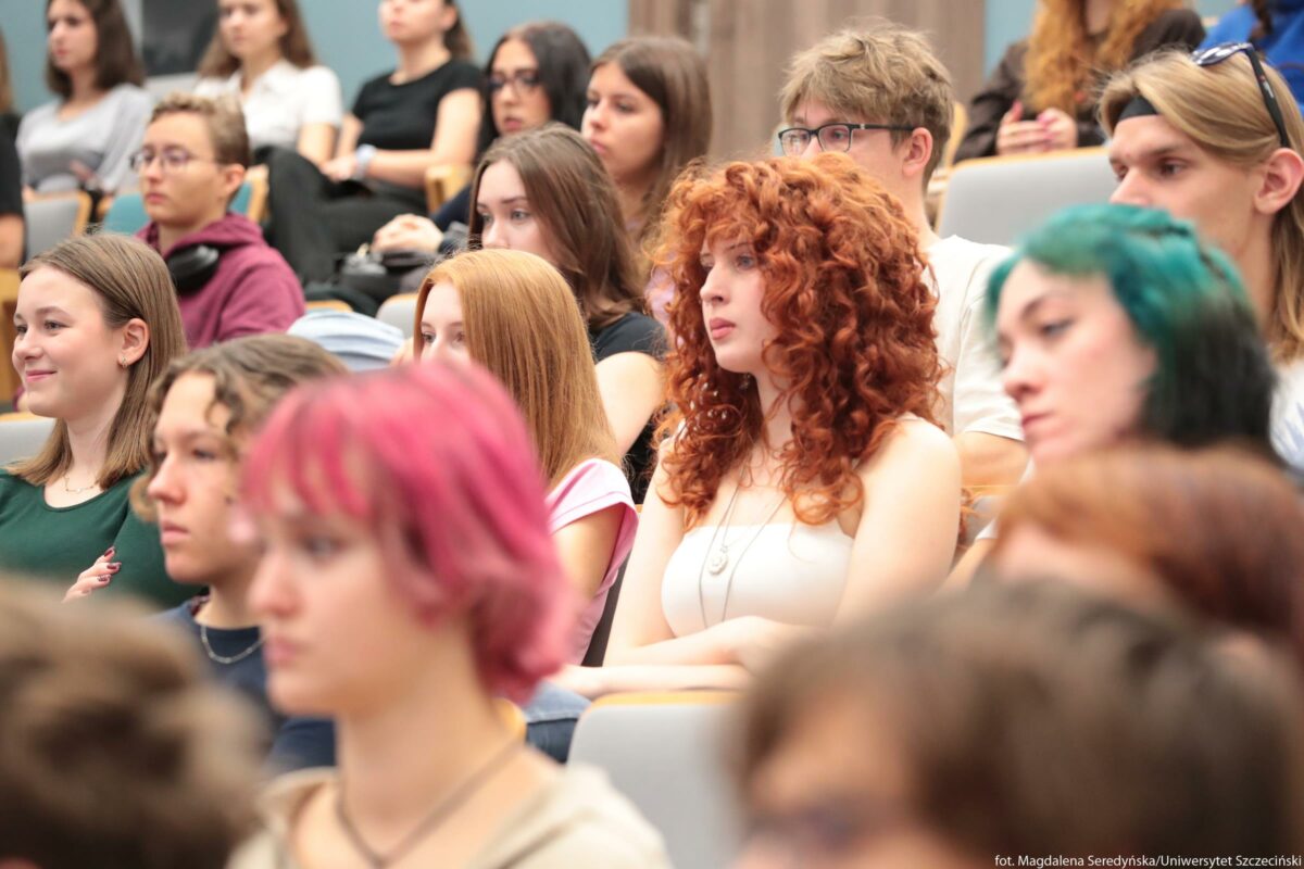 Grupa młodych osób siedzących w sali wykładowej. Widać kilkanaście studentek i studentów siedzących w rzędach na krzesłach z oparciami, skierowanych w jedną stronę, jak podczas wykładu lub spotkania. Najbardziej wyraźna jest osoba na środku kadru z bardzo kręconymi, długimi rudymi włosami, jasną skórą i białym topem na ramiączkach; ma skrzyżowane ręce na brzuchu i spokojny, skupiony wyraz twarzy. Po jej lewej stronie siedzi osoba z prostymi, długimi rudymi włosami, w czarnej koszulce, lekko pochylona do przodu. Z prawej strony widoczna jest osoba z włosami w odcieniach zieleni i turkusu, opadającymi na jedną stronę twarzy, z delikatnym makijażem, patrząca w tę samą stronę co reszta. Na pierwszym planie, bliżej obiektywu, znajduje się osoba z krótkimi, różowymi włosami, lekko rozmazana przez małą głębię ostrości, co podkreśla skupienie na środkowej części sali. W lewym dolnym rogu siedzi osoba w zielonej koszulce, lekko uśmiechnięta, o jasnej skórze i prostych włosach do ramion. W tle widać kolejne rzędy siedzeń zajęte przez osoby o zróżnicowanych fryzurach i kolorach włosów: od jasnego brązu i blond po ciemne i rudoczerwone; część ma okulary, część ma słuchawki zawieszone na szyi. Wszyscy wyglądają na nastolatków lub młodych dorosłych, skupionych na wydarzeniu odbywającym się poza kadrem. Tło jest jasne i neutralne, z fragmentami błękitnych i szarych ścian, co sugeruje nowoczesną salę uniwersytecką.