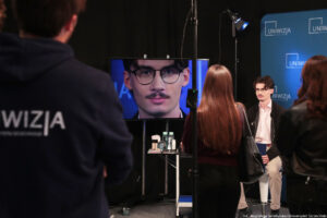 Na pierwszym planie trzy stojące tyłem osoby, w tym po lewej mężczyzna z białym napisem Uniwizja na ciemnej bluzie. Po prawej młody mężczyzna siedzący na hokerze (przodem do kadru), a za nim granatowa ścianka z niebieskimi logami Uniwizji. Po lewej ten sam mężczyzna na dużym ekranie.