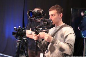 Ujęcie bokiem. Młody mężczyzna trzymający kamerę i patrzący na zarejestrowany przez nią obraz. W tle kamera na niskim statywie i kamerzysta (zasłonięty przez kamerę).