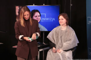 Studentka siedząca na hokerze w założonej jasnej pelerynie (ujęcie do kolan). Po lewej kobieta przygotowująca osoby do występu na wizji, która nalewa płyn na bawełniany płatek. W tle duży ekran, na którym widać studentkę.