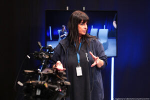 Studio telewizyjne. Beata Mikołajewska-Wieczorek – dyrektorka Centrum Edukacji Medialnej i Interaktywności. Po lewej stronie kamery. W tle duży ekran z rejestrowanym obrazem.