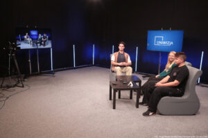 Studio telewizyjne. Ciemne tło. Po lewej w rogu kamera na statywie i ekran z rejestrowanym obrazem. Po prawej dwa niskie kwadratowe czarne stoliki i trzy szare fotele (jeden dla prowadzącego – na wprost, dwa dla gości – bokiem). Za ludźmi ekran z granatowym tłem i białym logiem Uniwizja.