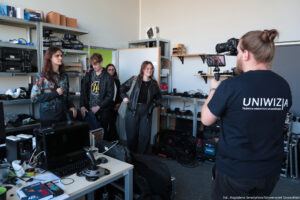 Pokój montażowy, do którego wchodzi czworo studentów (dwie studentki i dwóch studentów). Dookoła metalowe półki ze sprzętem typu, kamery, kable, skrzynki. Po prawej (tyłem) kamerzysta w ciemnej koszulce z białym logiem Uniwizji, nagrywający uczestników Dni Adaptacyjnych.