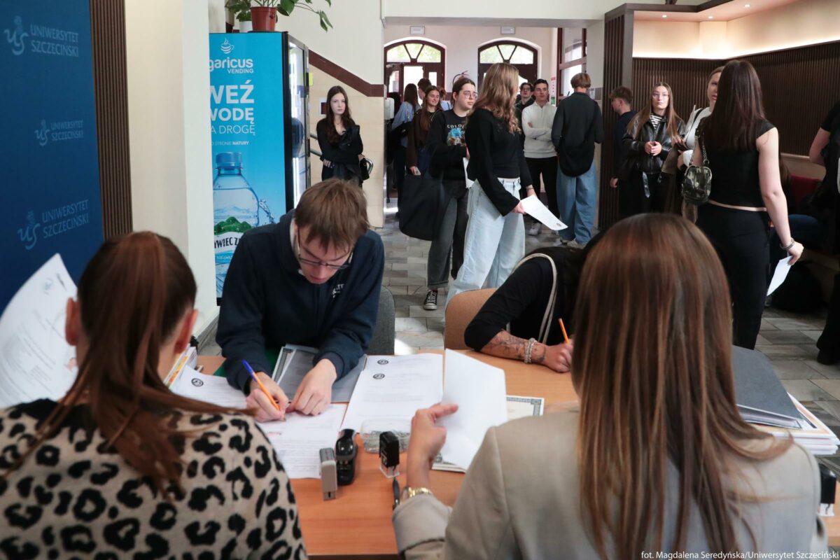 Pomieszczenie w budynku uczelni, w którym odbywa się obsługa studentów przy stoliku. Na pierwszym planie jest drewniany stół, przy którym siedzą trzy osoby odwrócone częściowo tyłem do obiektywu; przed nimi leżą pliki dokumentów, pieczątki i długopisy. Dwie osoby pochylają się nad formularzami i coś wypełniają, trzecia trzyma kilka kartek w dłoni. W tle widać korytarz uczelniany pełen młodych osób, większość w casualowych ubraniach; część trzyma dokumenty, część rozmawia, kilka osób stoi w kolejce. Po lewej stronie kadru znajduje się niebieska ścianka z białym napisem “Uniwersytet Szczeciński” i logotypem uczelni, obok stoi automat z napojami z dużą reklamą butelki wody w kolorze niebieskim. Na końcu korytarza są przeszklone drzwi wejściowe z łukowymi oknami, przez które wpada dzienne światło. Atmosfera przypomina dzień rejestracji lub spraw organizacyjnych na uniwersytecie.