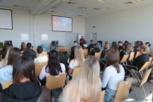 Sala wykładowa (ujęcie bokiem). Około 30-osobowa grupa studentów siedzących na drewnianych krzesłach (tyłem do kadru). W tle biurko z komputerem i przedstawicielka Akademickiego Biura Karier prowadząca Dni Adaptacyjne. U góry ekran z napisem #FunRunUS.