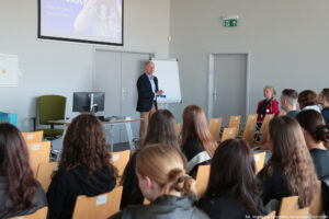 Sala wykładowa (ujęcie bokiem). Widok na 10-osobowę grupę studentów siedzących na drewnianych krzesłach (tyłem do kadru). W tle biurko z komputerem i przedstawiciel uczelni prowadzący Dni Adaptacyjne. U góry ekran z napisem Stacja motywacja, KampUS Kariera.