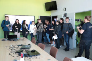 Pokój konferencyjny. Z przodu długi stół z krzesłami wzdłuż niego. Na stole laptopy i kable. W tle około 10-osobowa grupa studentów. W prawym rogu bokiem kamerzysta w ciemnej koszulce z białym logiem Uniwizji, nagrywający uczestników Dni Adaptacyjnych.