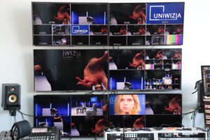 Pokój transmisyjny. Sześć ekranów zawieszonych na ścianie w dwóch pionowych rzędach, na których wyświetlają się rejestrowane obrazy. Każdy ekrany podzielony na cztery różne widoki. Na dole konsole telewizyjne z przyciskami.